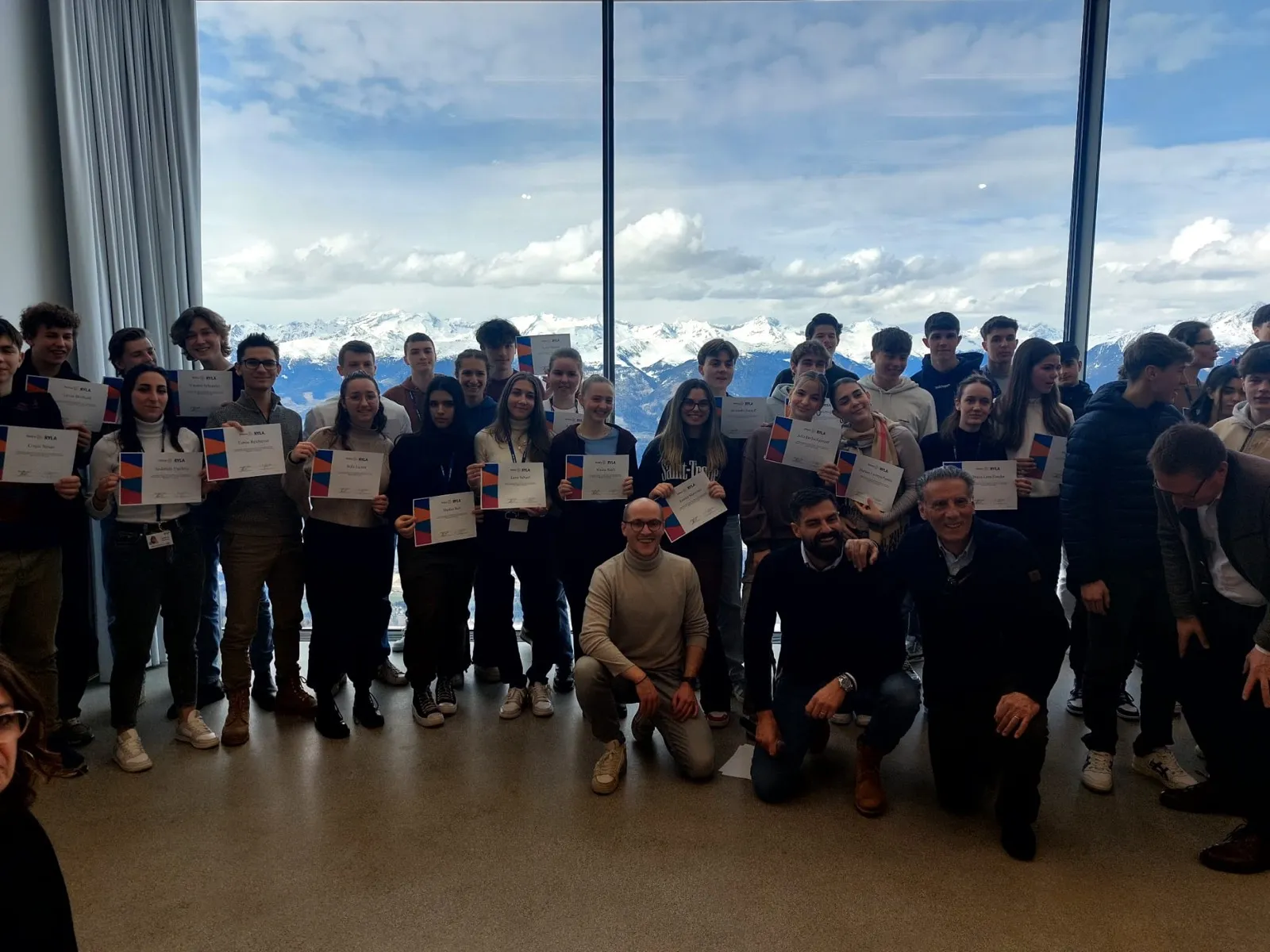Large group of participants holding certificates in front of a panoramic window overlooking snow-covered mountains, three organisers kneeling in the front row