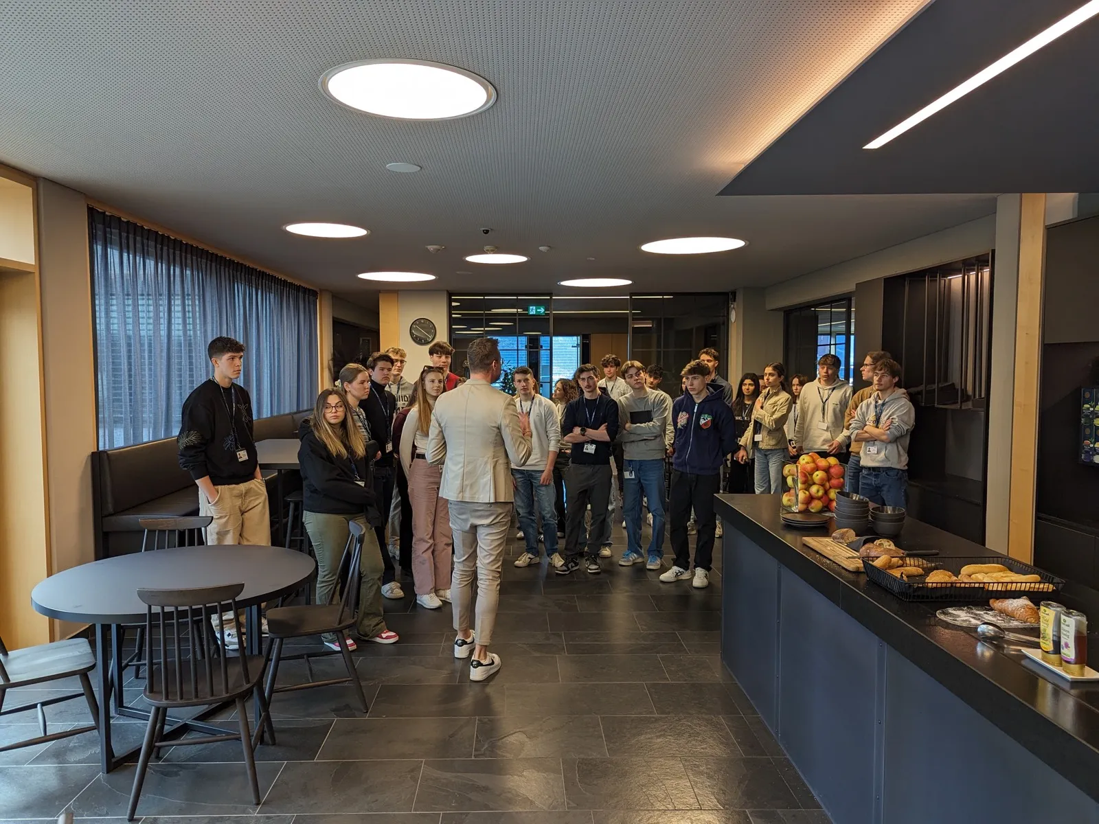 A group of teenagers standing around a speaker in a modern company lounge, with a fruit bowl and pastry basket on the counter beside them