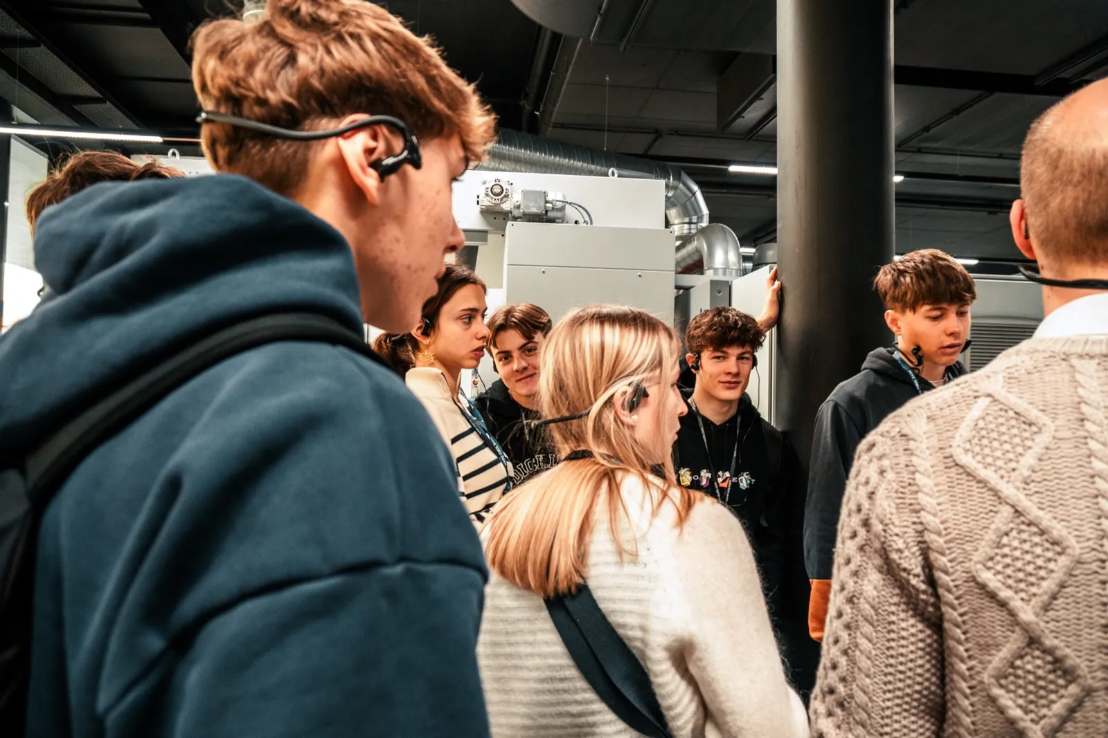 Participants wearing tour headsets crowded around an industrial machine on a shop floor, one listener with headphones in the foreground