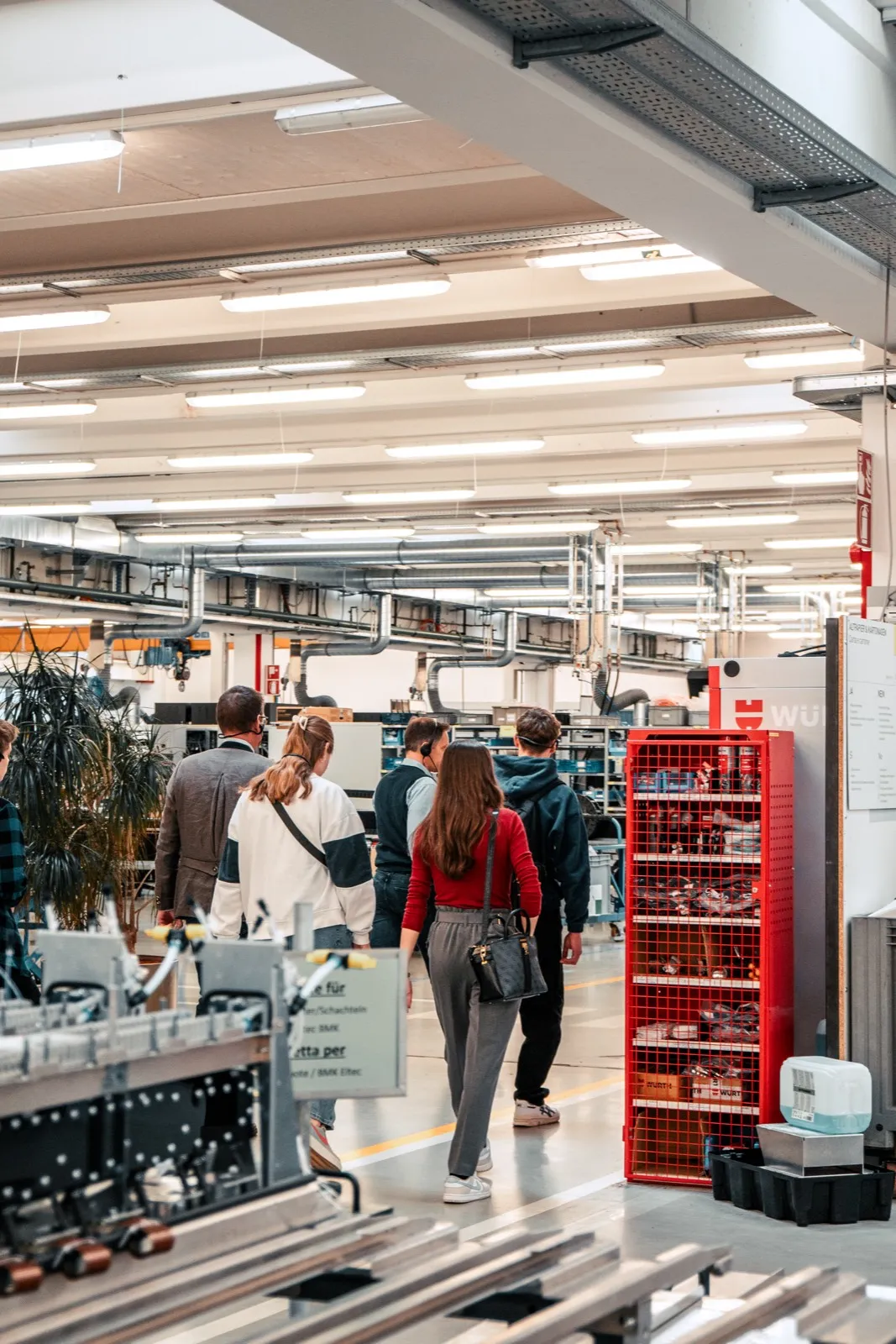 A small group of visitors walking through a lit workshop hall past a red parts shelf and machinery, cable trays overhead