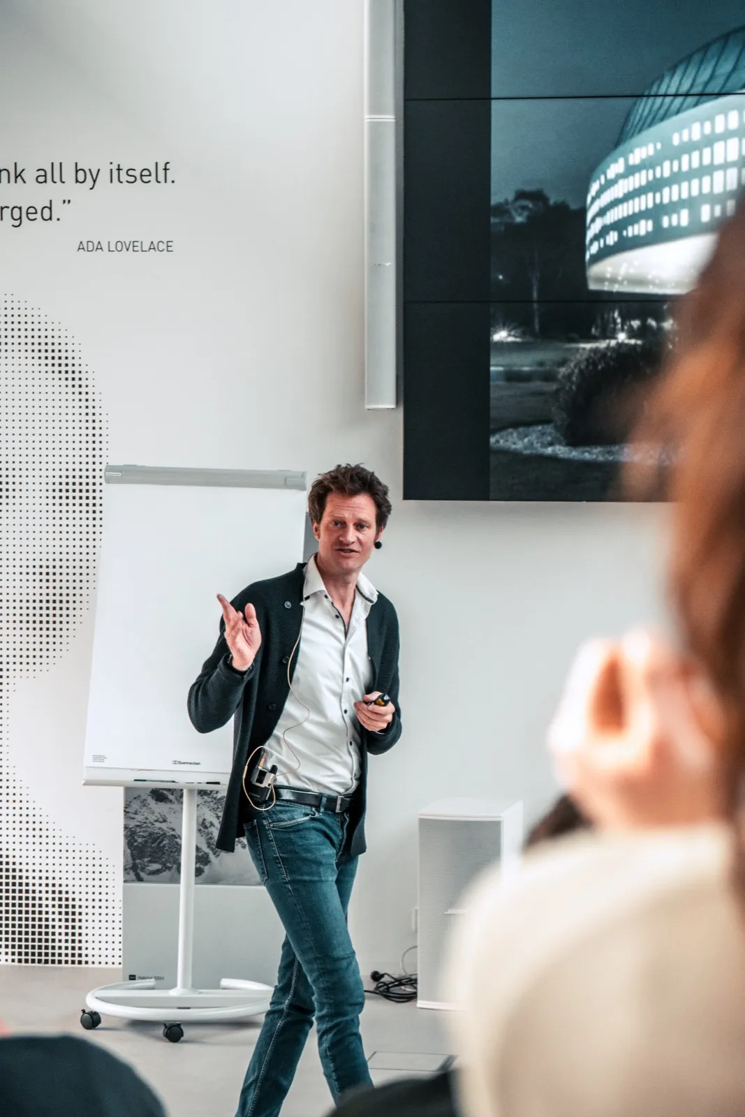 A speaker gesturing in front of a wall panel bearing a printed quotation, next to a flipchart on a wheeled stand and a large photographic print