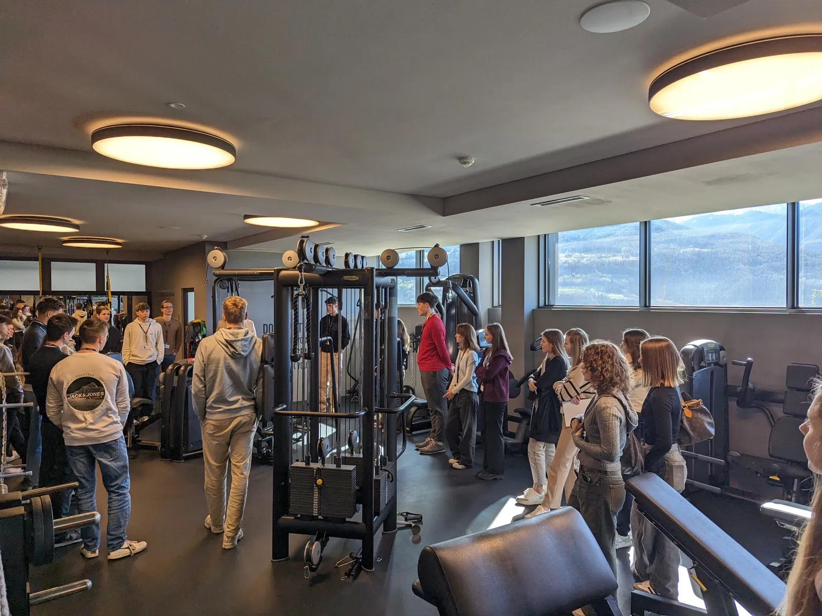 Participants in a modern fitness studio around cable-machine equipment, an instructor addressing them beside a large window overlooking mountains outside