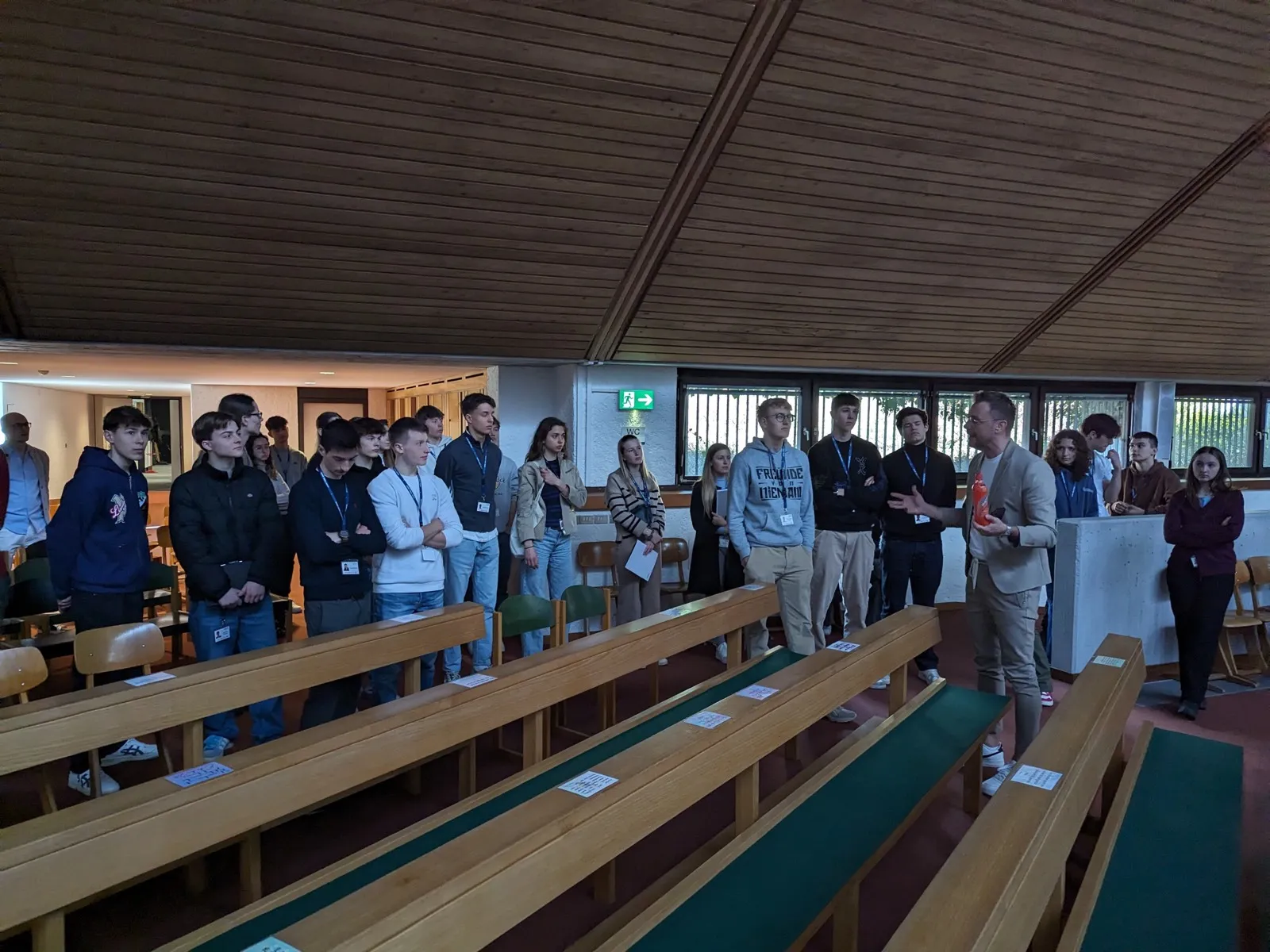 A group of participants standing between wooden pews in a modern chapel, listening to an organiser in a rust-coloured jacket, with programme sheets on the benches