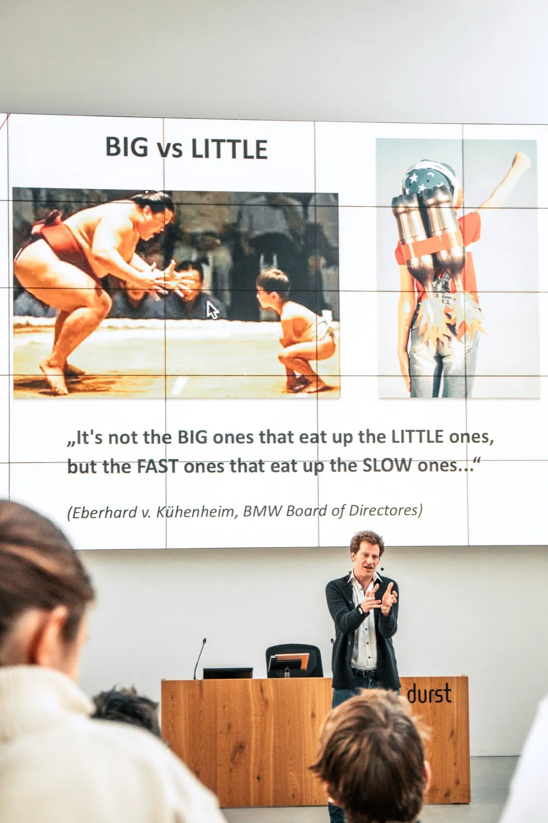 A speaker gesturing in front of a large video wall showing a sumo wrestler on the left and a jet-pack figure on the right, a wooden Durst lectern in front