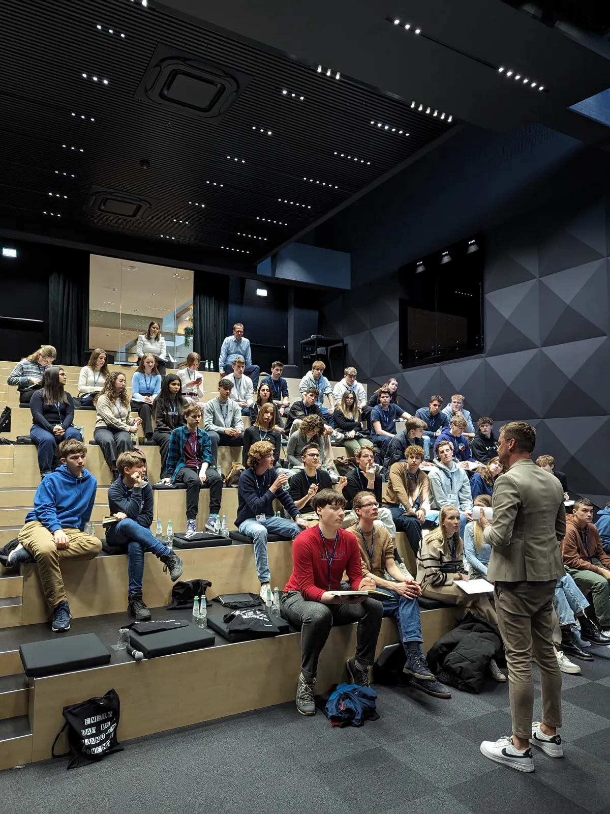 A tiered wooden auditorium of seated teenagers with an instructor in a beige suit standing at the front and one student in a red hoodie in the middle row