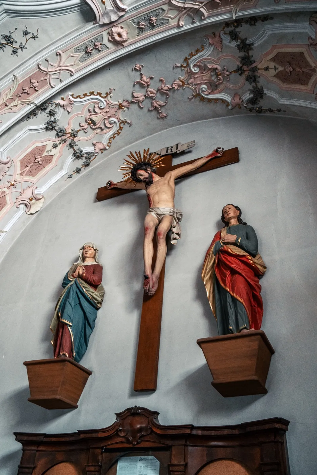 A baroque wooden crucifix on a chapel wall with ornate stucco and two flanking painted statues, one holding a red-draped scripture