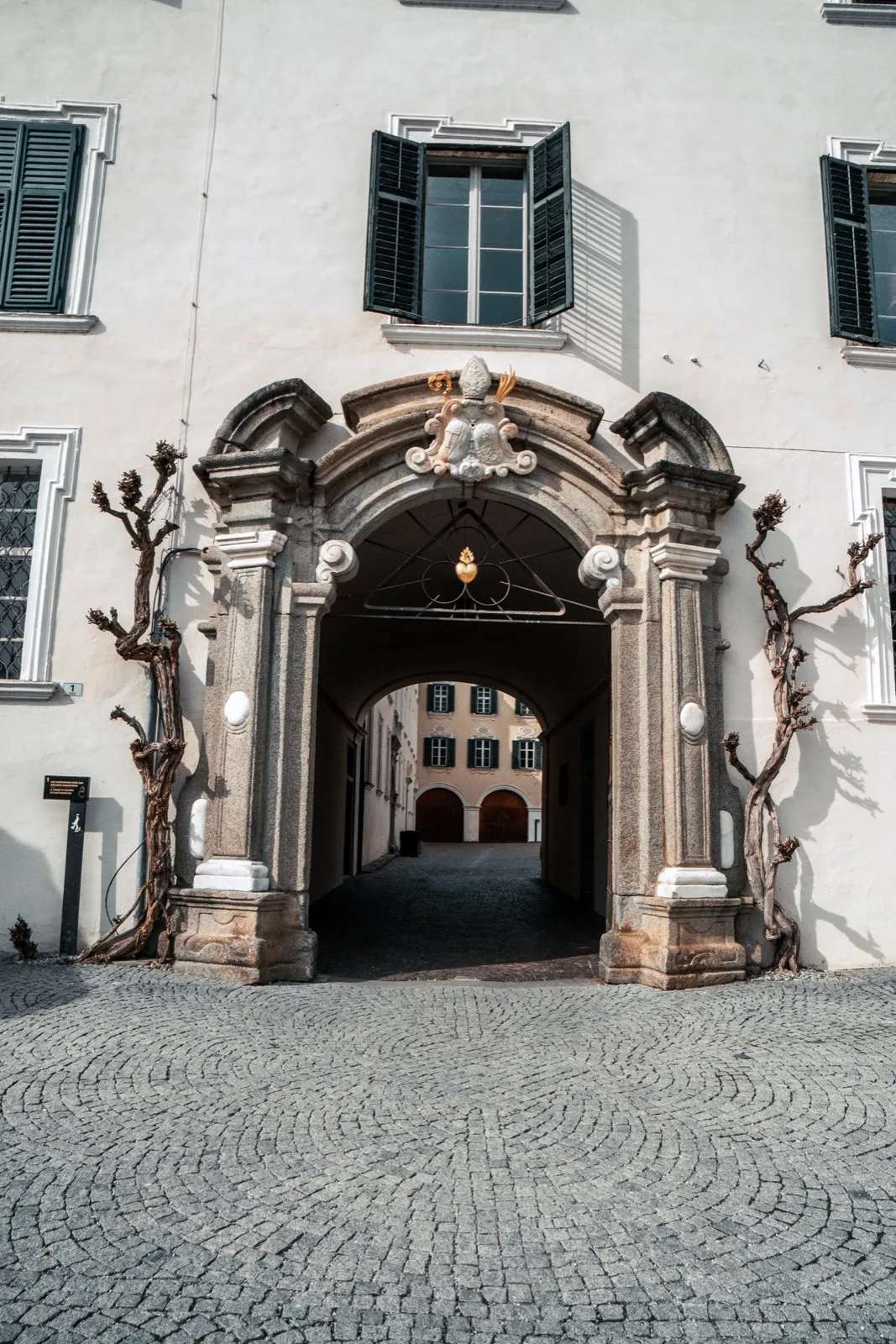 A baroque stone portal with paired columns and a coat of arms leading into a cobbled inner courtyard, pruned vines climbing on the wall beside it
