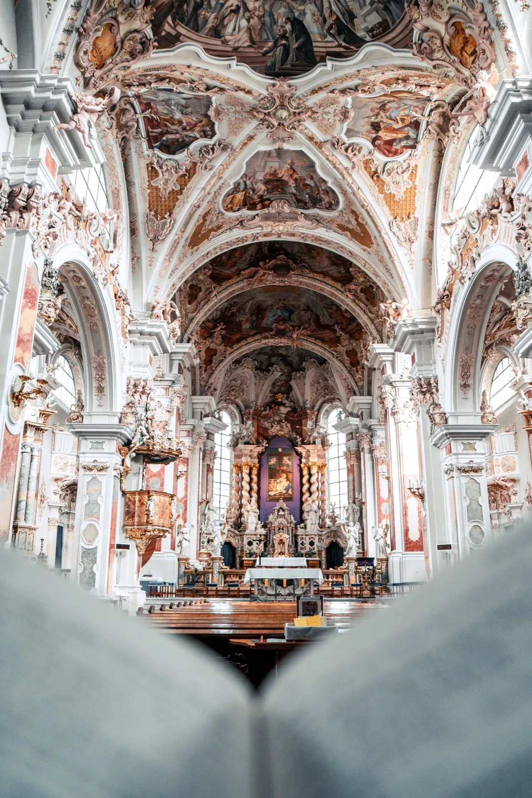 A baroque monastery church seen from the pews with a frescoed vaulted ceiling, an ornate marble altar with twisted columns, and an open hymnal in the foreground