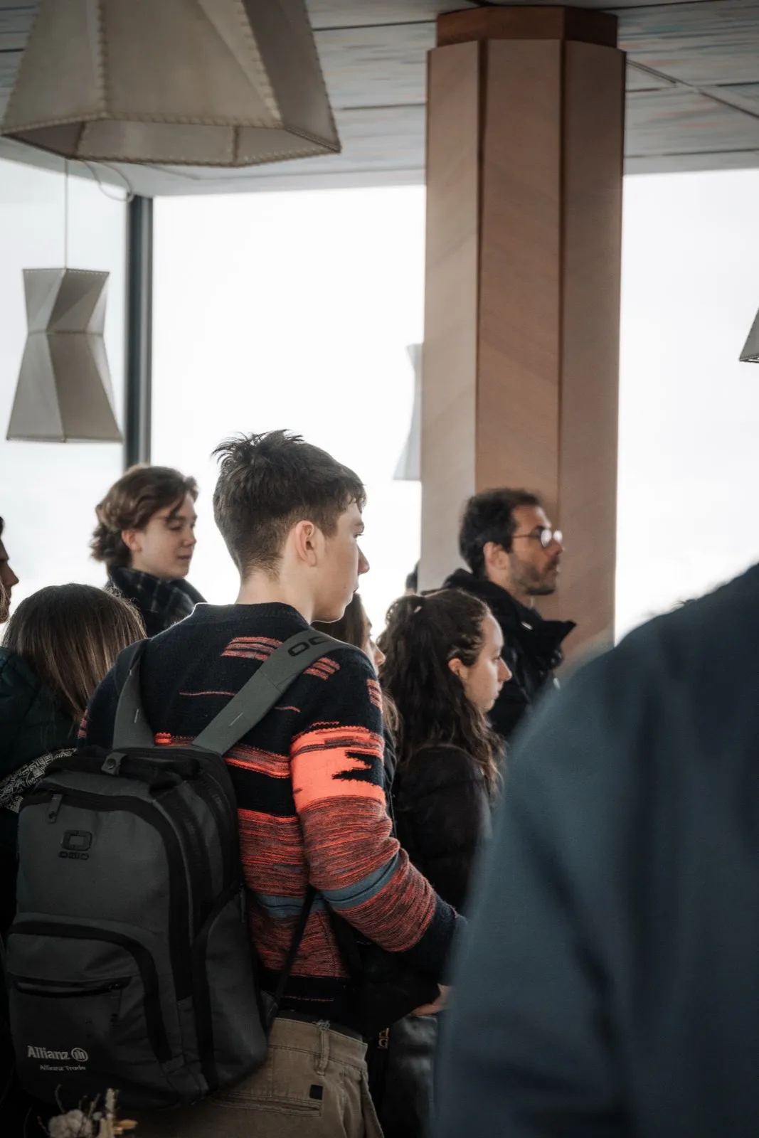 Teenagers standing in a mountain-top restaurant with lantern-shaped lamps, one wearing a backpack, waiting close to a large panoramic window