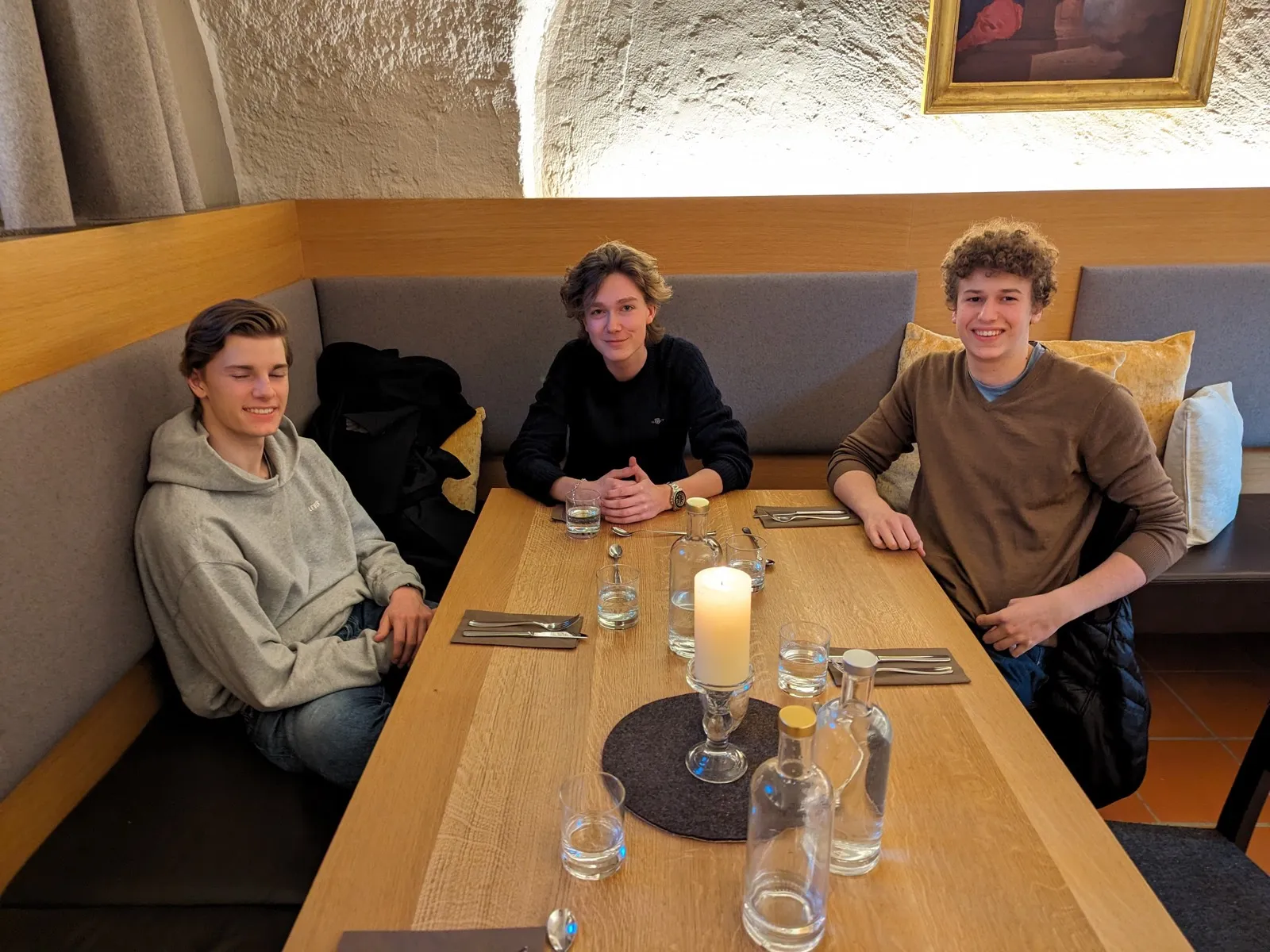Three young men seated at a wooden restaurant booth with a lit candle, water carafes, and small glasses arranged on the table