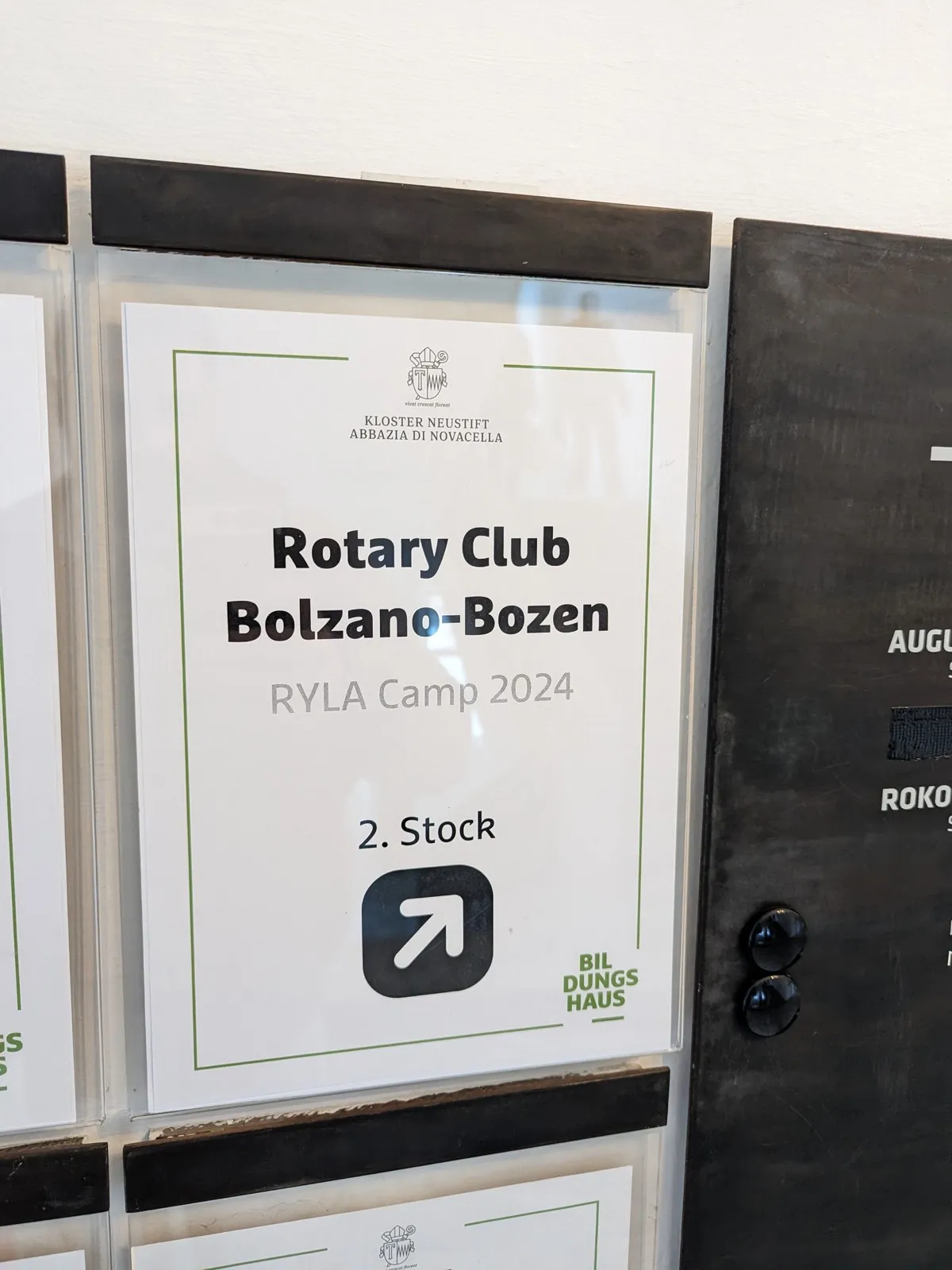 A printed door sign inside a plastic sleeve reading Rotary Club Bolzano-Bozen RYLA Camp with an arrow directing visitors to the second floor