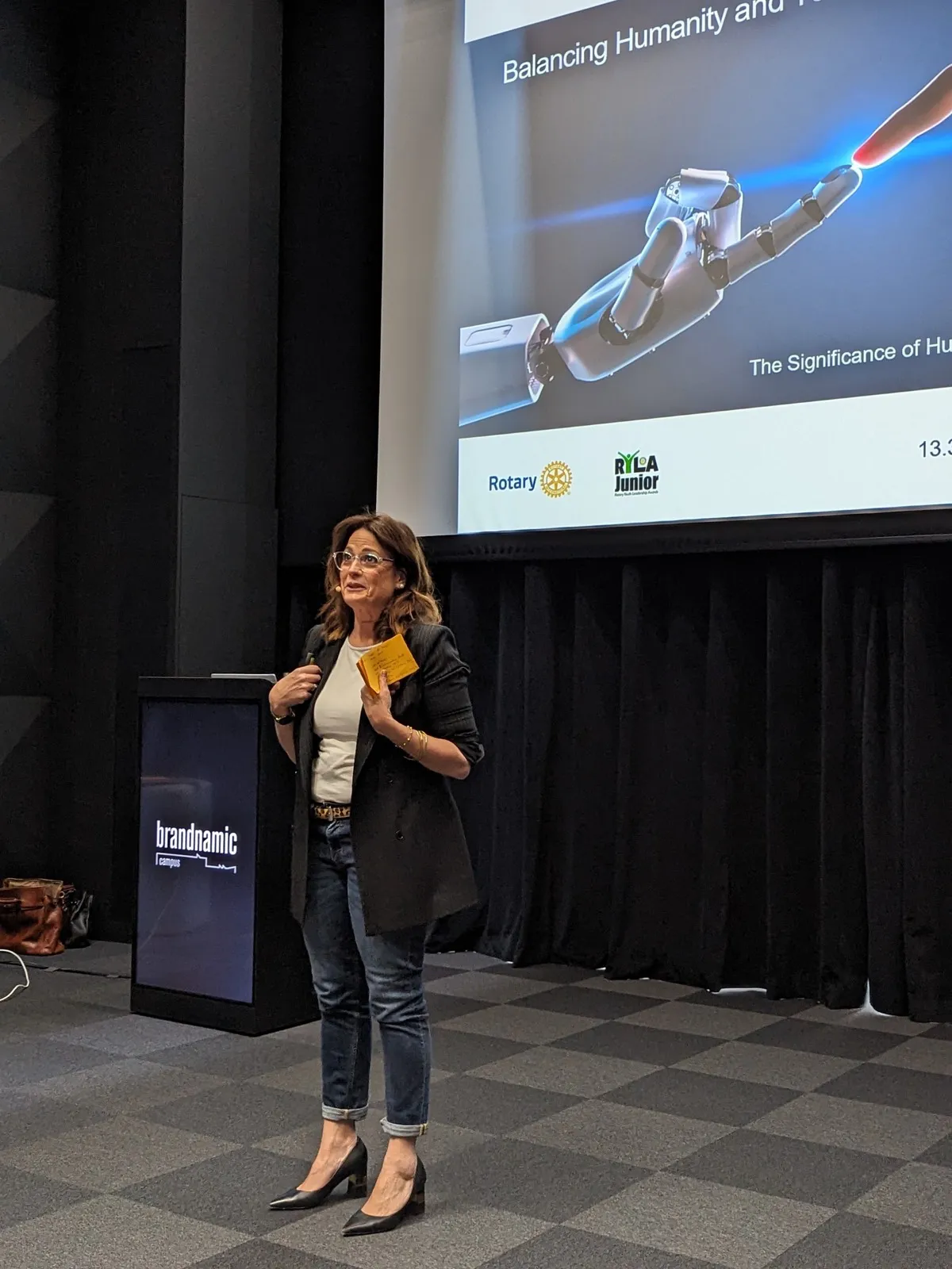 A woman in dark blazer and jeans speaking at a lectern beside a screen showing a robotic hand touching a human finger