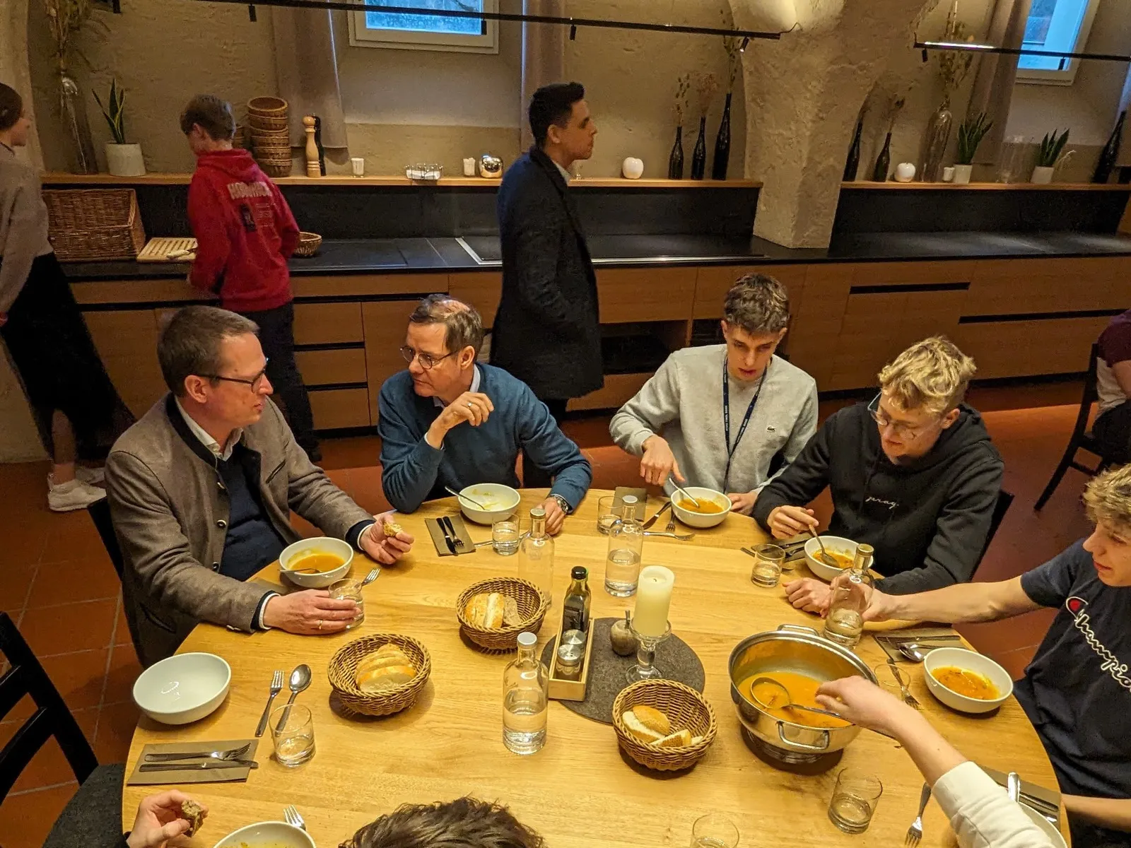 A group gathered around a round wooden table sharing bowls of orange soup, bread baskets, and glass carafes inside a vaulted dining room