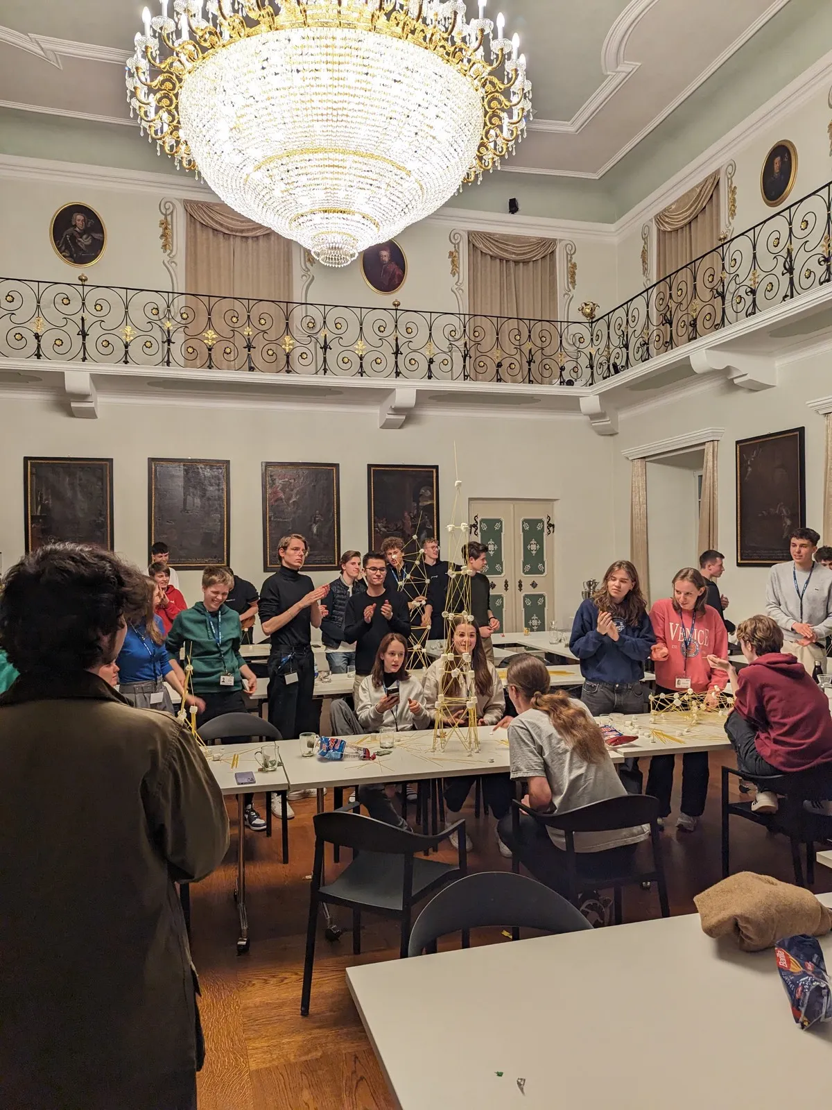 Teenagers building tall spaghetti and marshmallow towers on white tables beneath an ornate crystal chandelier in a portrait-lined hall