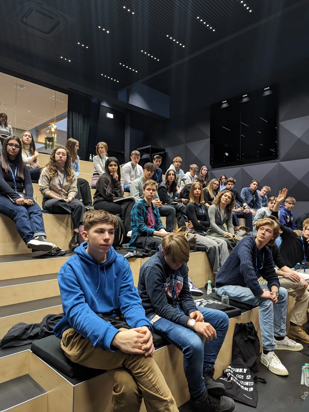 Rows of teenagers wearing lanyards seated on stepped wooden bleachers listening attentively inside a dark auditorium with wall-mounted screens