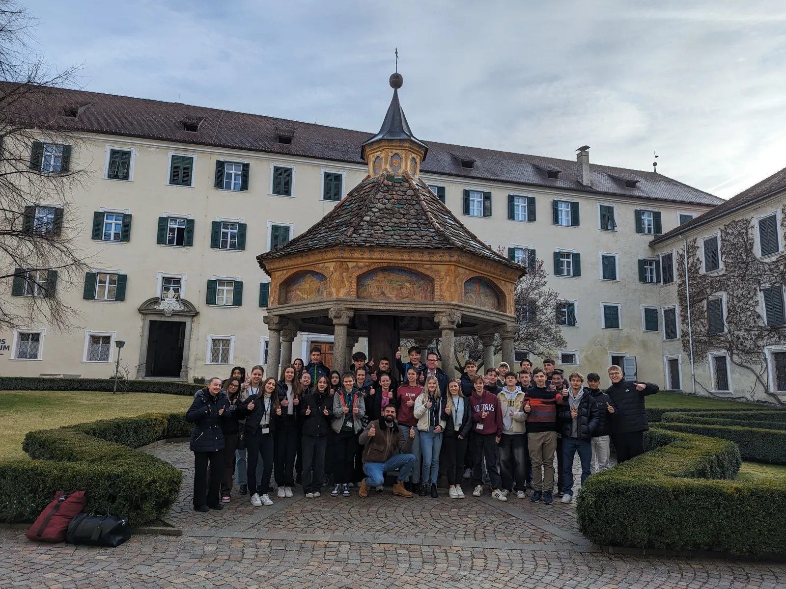 A large group posing with thumbs raised in front of a baroque courtyard pavilion framed by symmetrical hedges and a long shuttered building