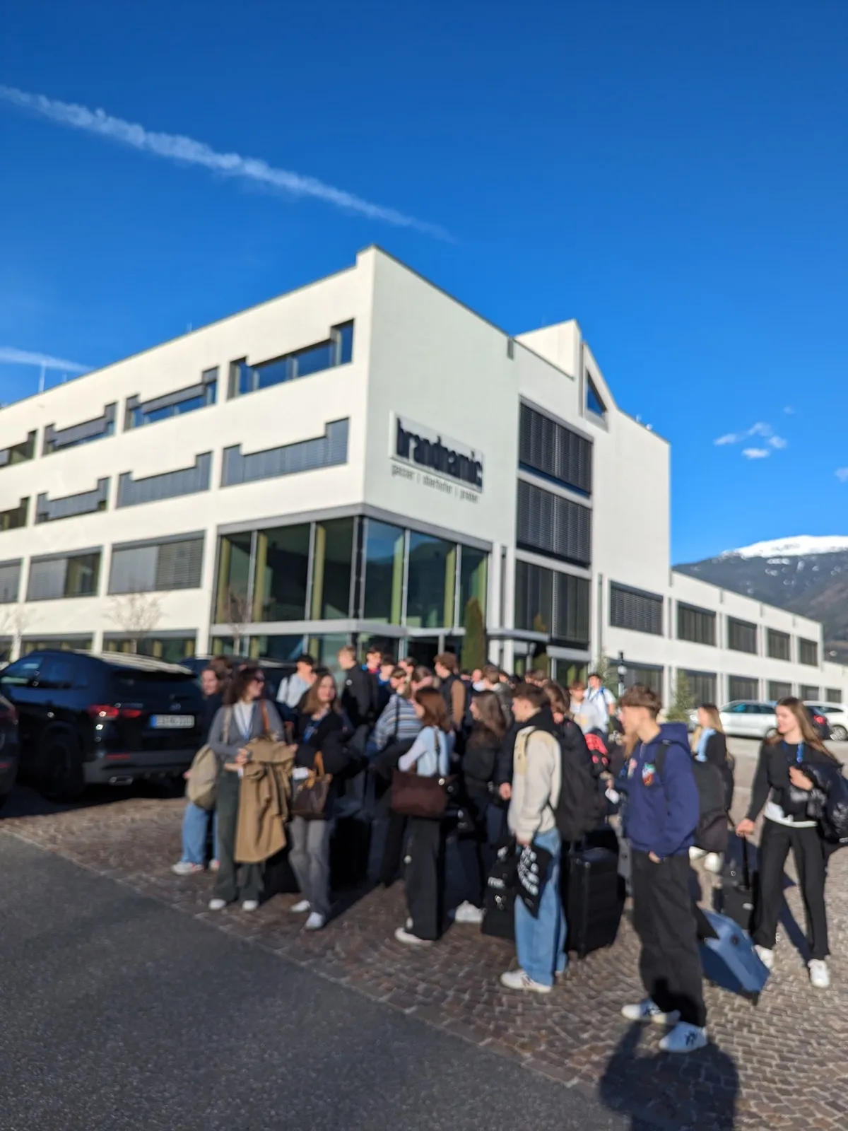 A crowd of teenagers carrying luggage and backpacks gathered outside the brandnamic office building under a bright blue sky with distant snowy peaks