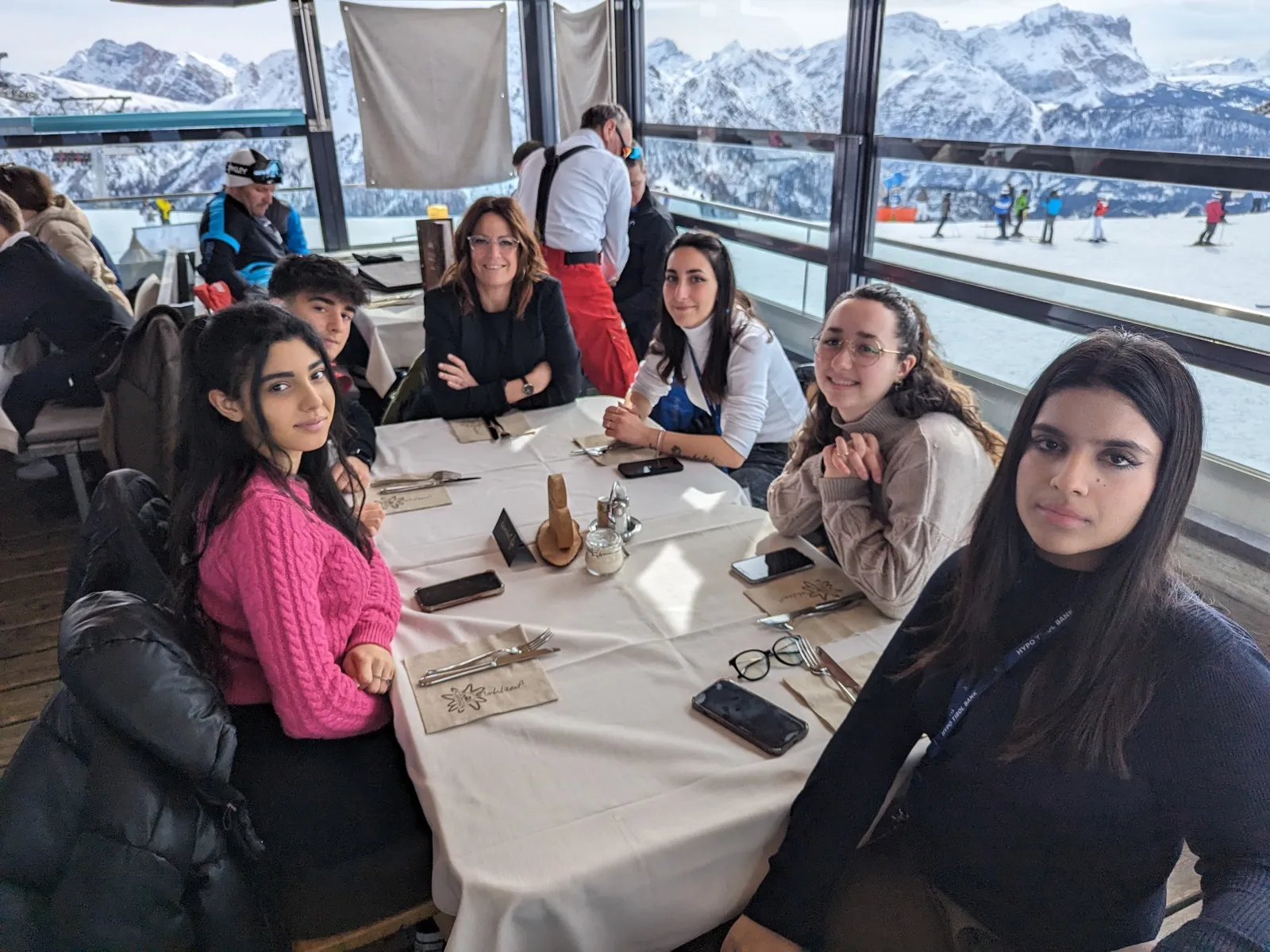 Six people seated around a linen-covered table inside a panoramic mountain restaurant with snowy Dolomite peaks visible through the windows