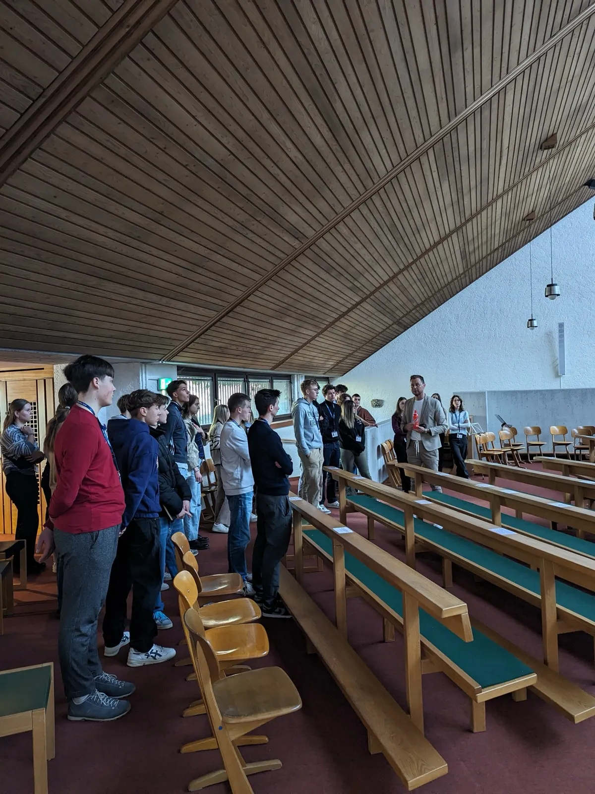 A group of participants standing between wooden pews inside a modern chapel, listening to a guide gesturing near the altar wall