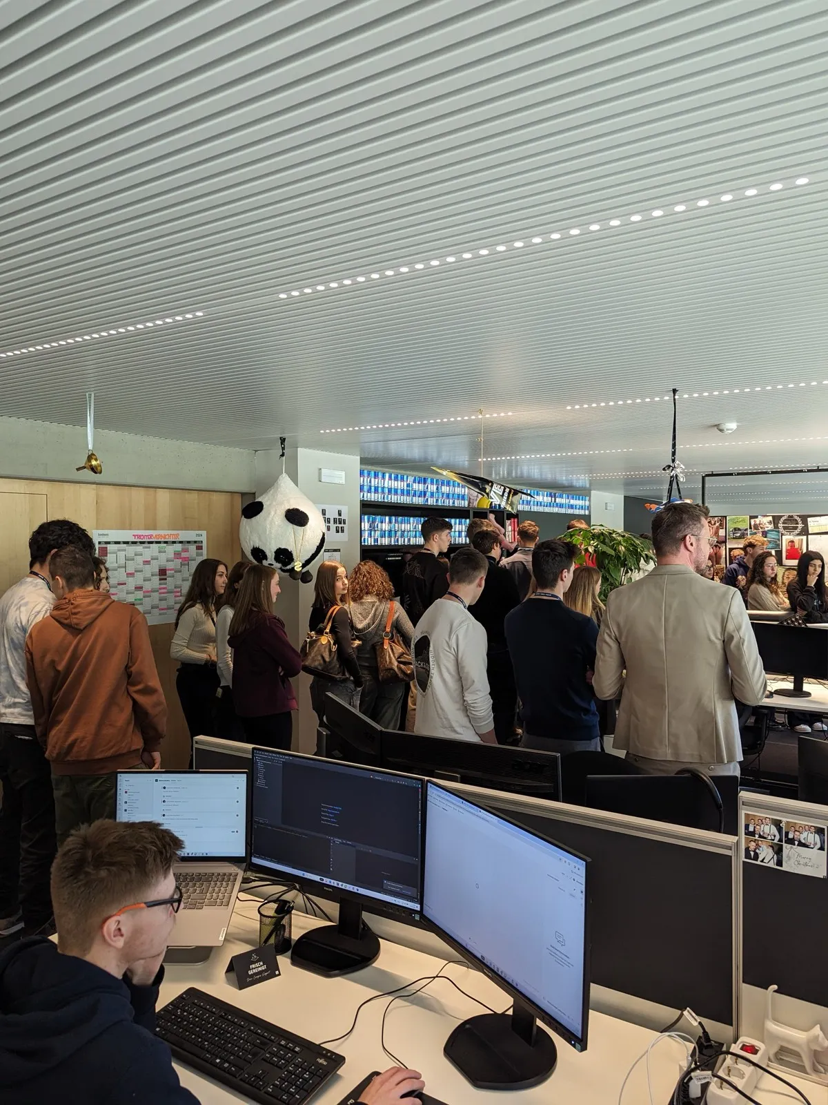 Participants crowding around desks in a company control room lined with screens, an organiser standing among them in a blazer
