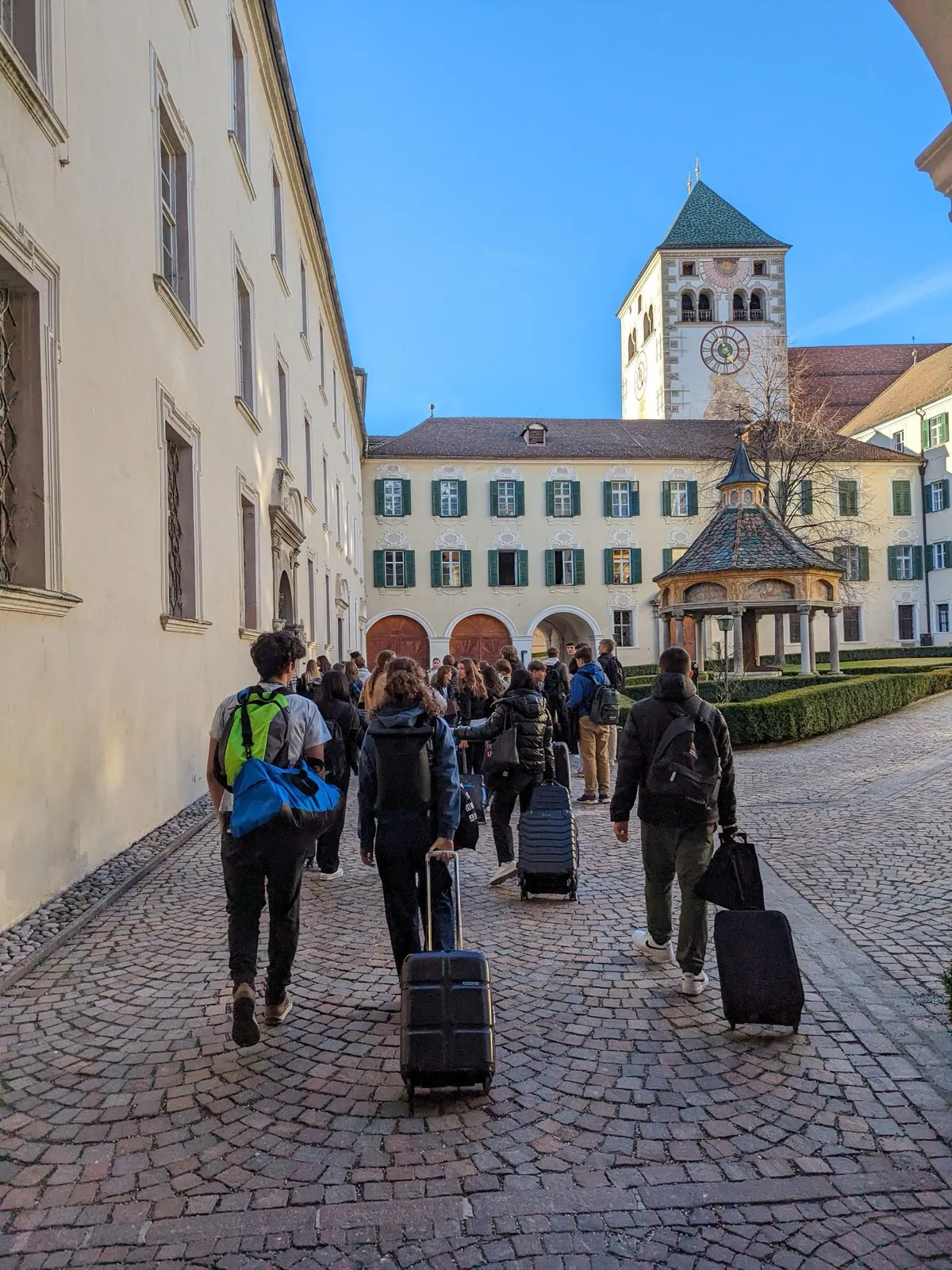A line of participants pulling suitcases through a cobbled monastery courtyard toward an arched gateway and a clock tower
