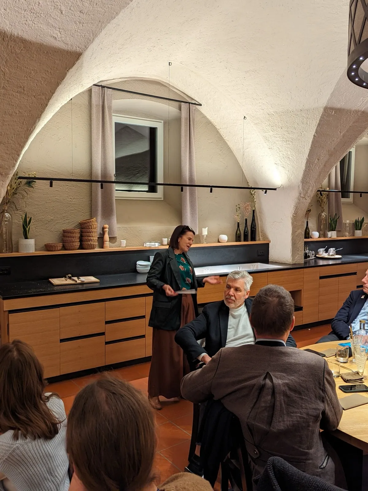 A speaker gesturing beside seated guests in a vaulted stone kitchen with open shelving and hanging curtains