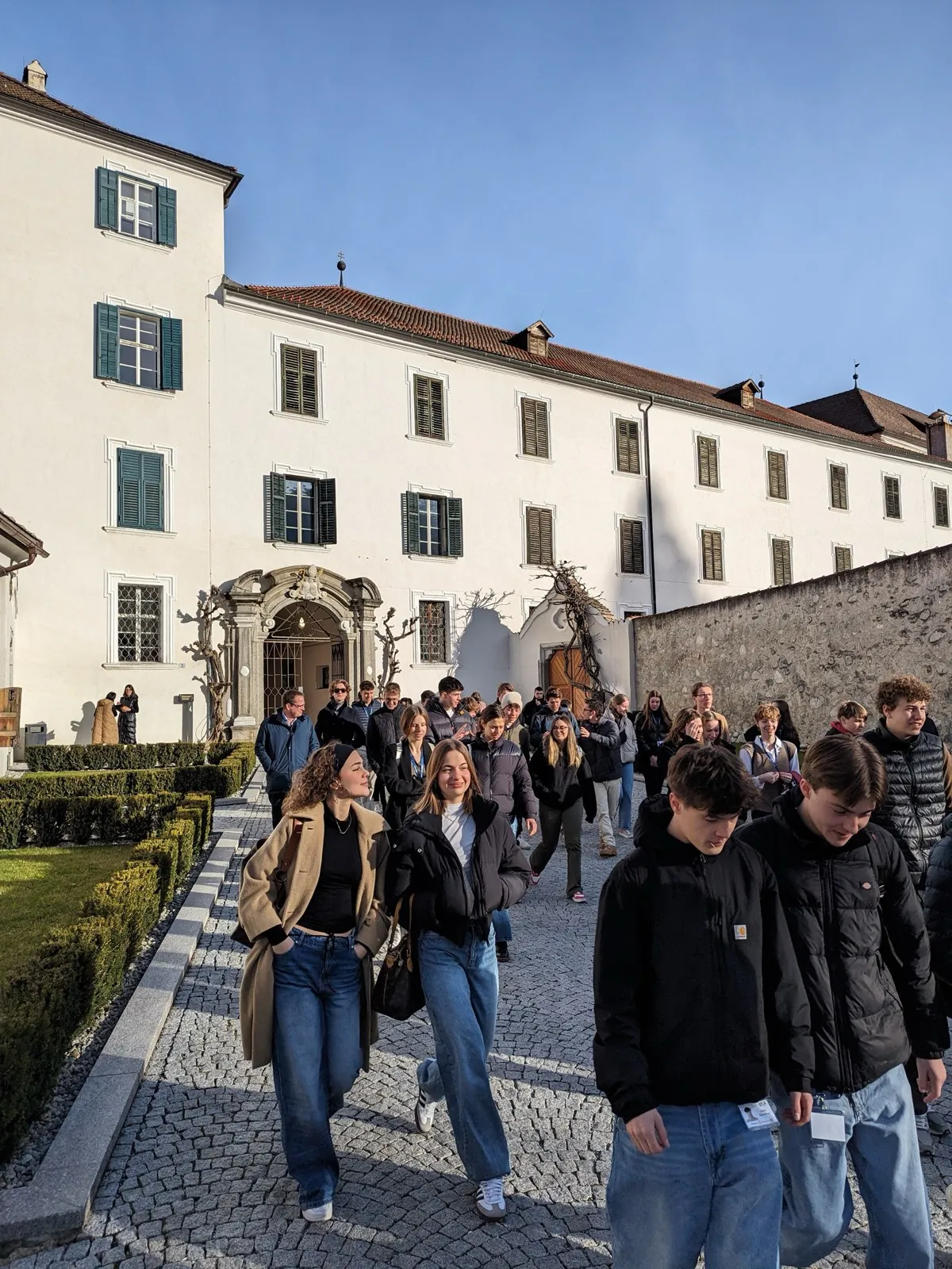 A group of participants walking through a sunlit monastery courtyard past a clipped hedge garden and a baroque gateway