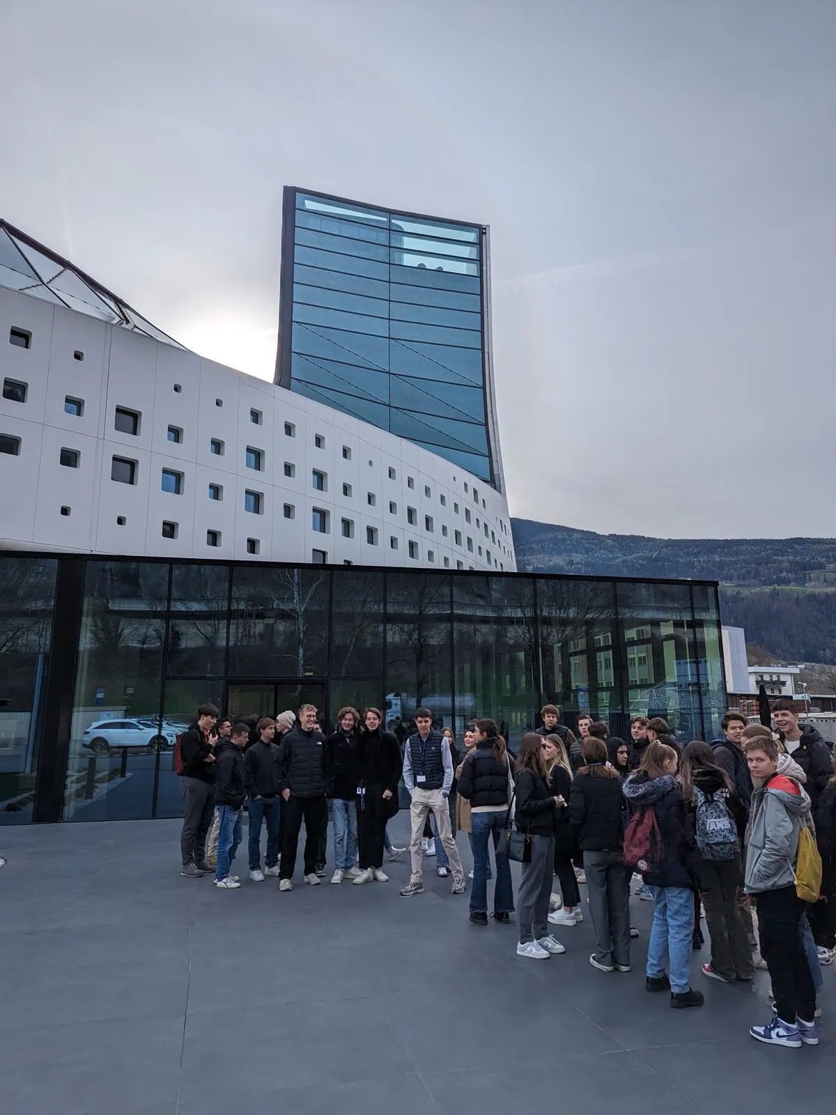 Participants gathered outside a modern glass-fronted building beside a tall glass tower, mountains rising behind under an overcast sky