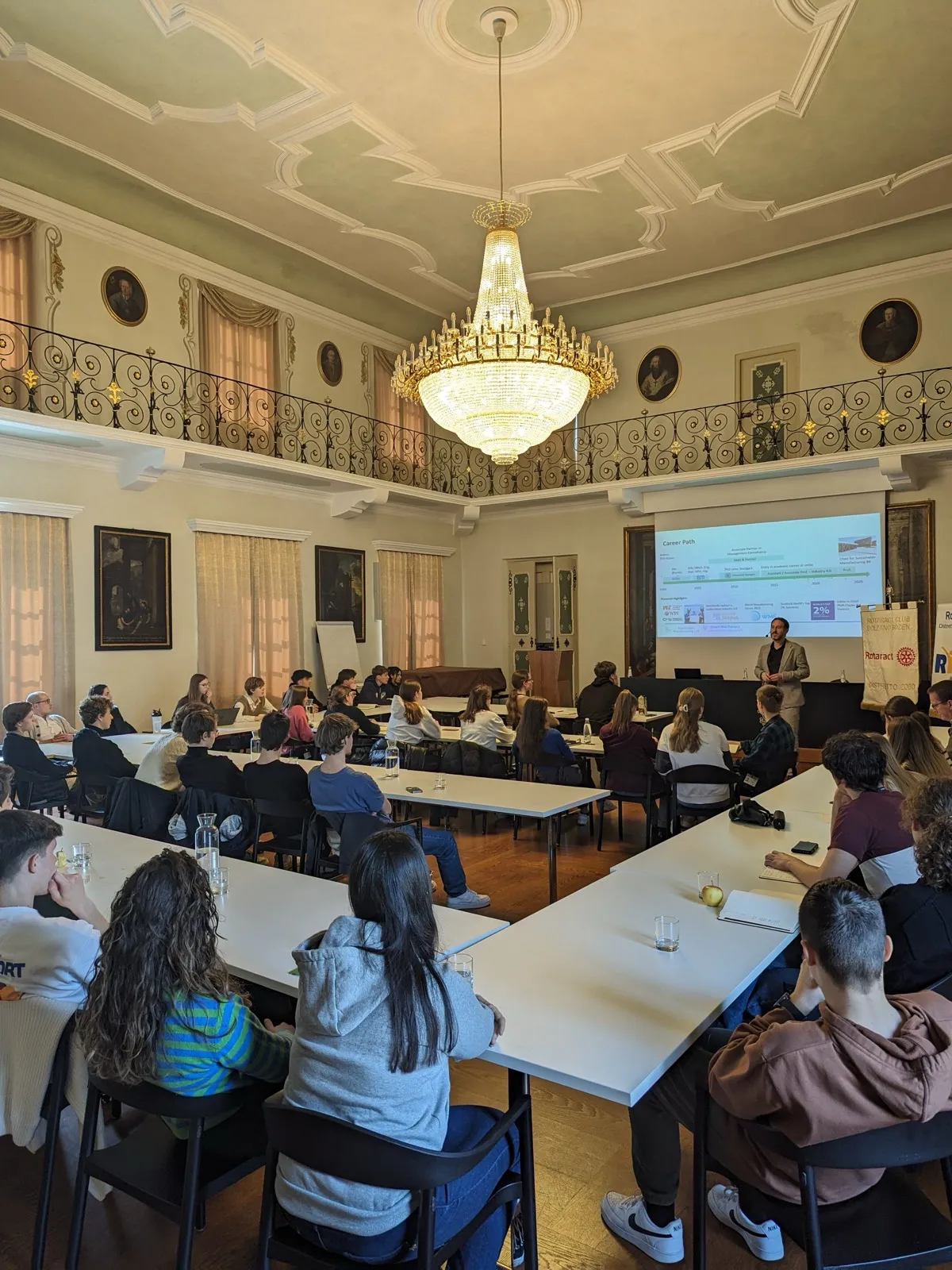 Participants seated at long tables in a chandelier-lit hall with a balcony gallery, facing a speaker beside a projected career timeline