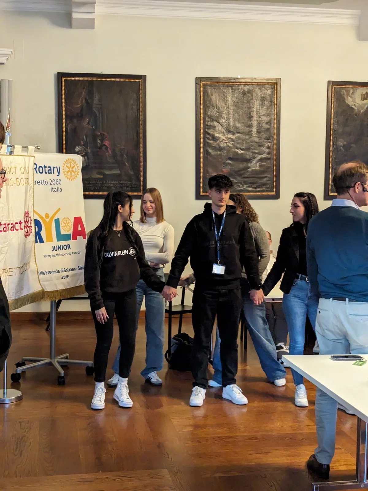 A line of participants holding hands during a team exercise in a portrait-lined hall, beside a RYLA banner and pennants