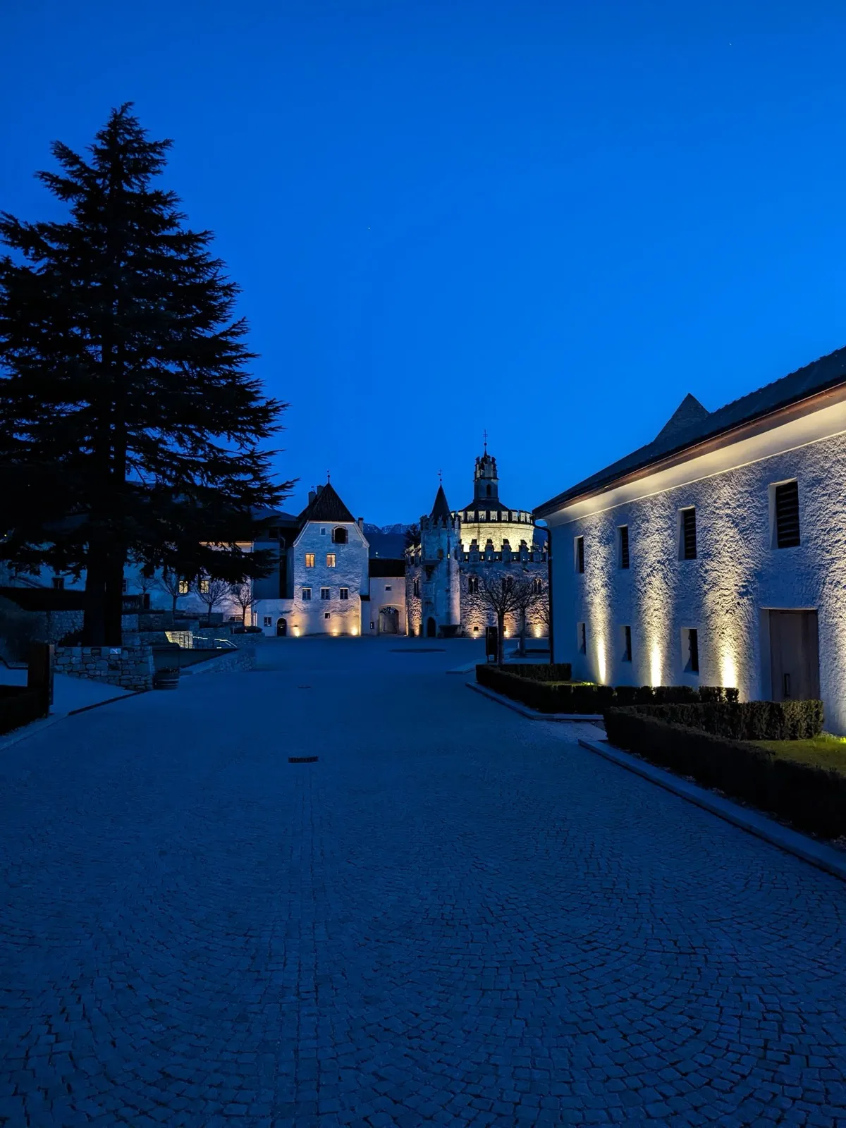 A floodlit monastery courtyard at dusk with a stone gate tower and dome glowing against a deep blue evening sky