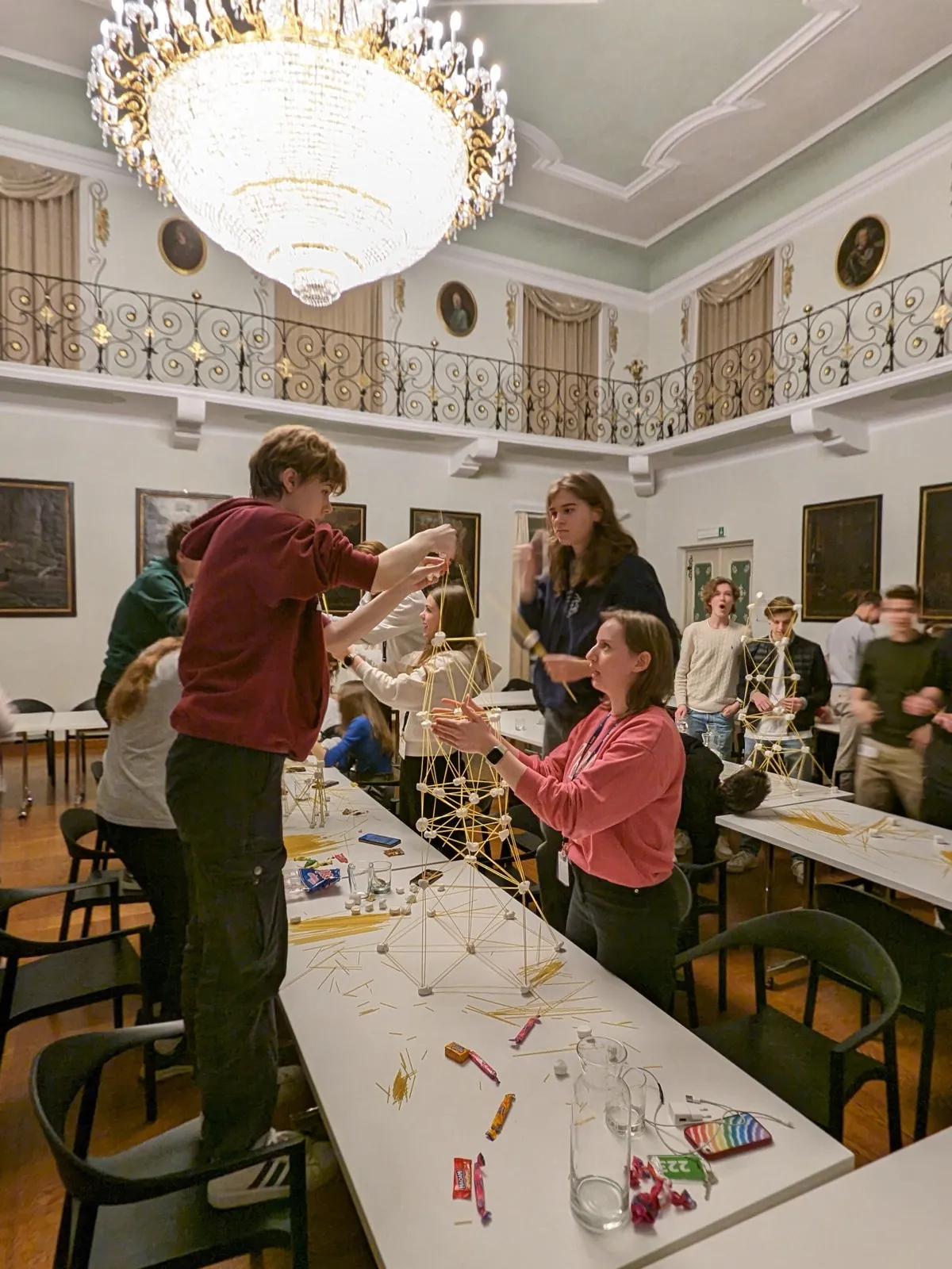 Participants building tall marshmallow and spaghetti towers at long tables beneath a crystal chandelier in a portrait-lined hall