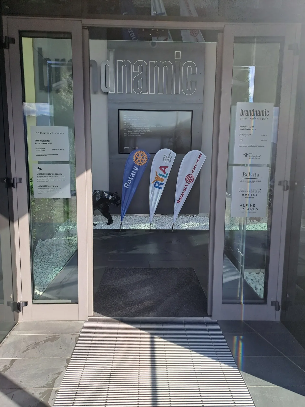 Glass entrance doors of an office building displaying flags for Rotary, Rotaract and RYLA next to Brandnamic signage and printed information sheets
