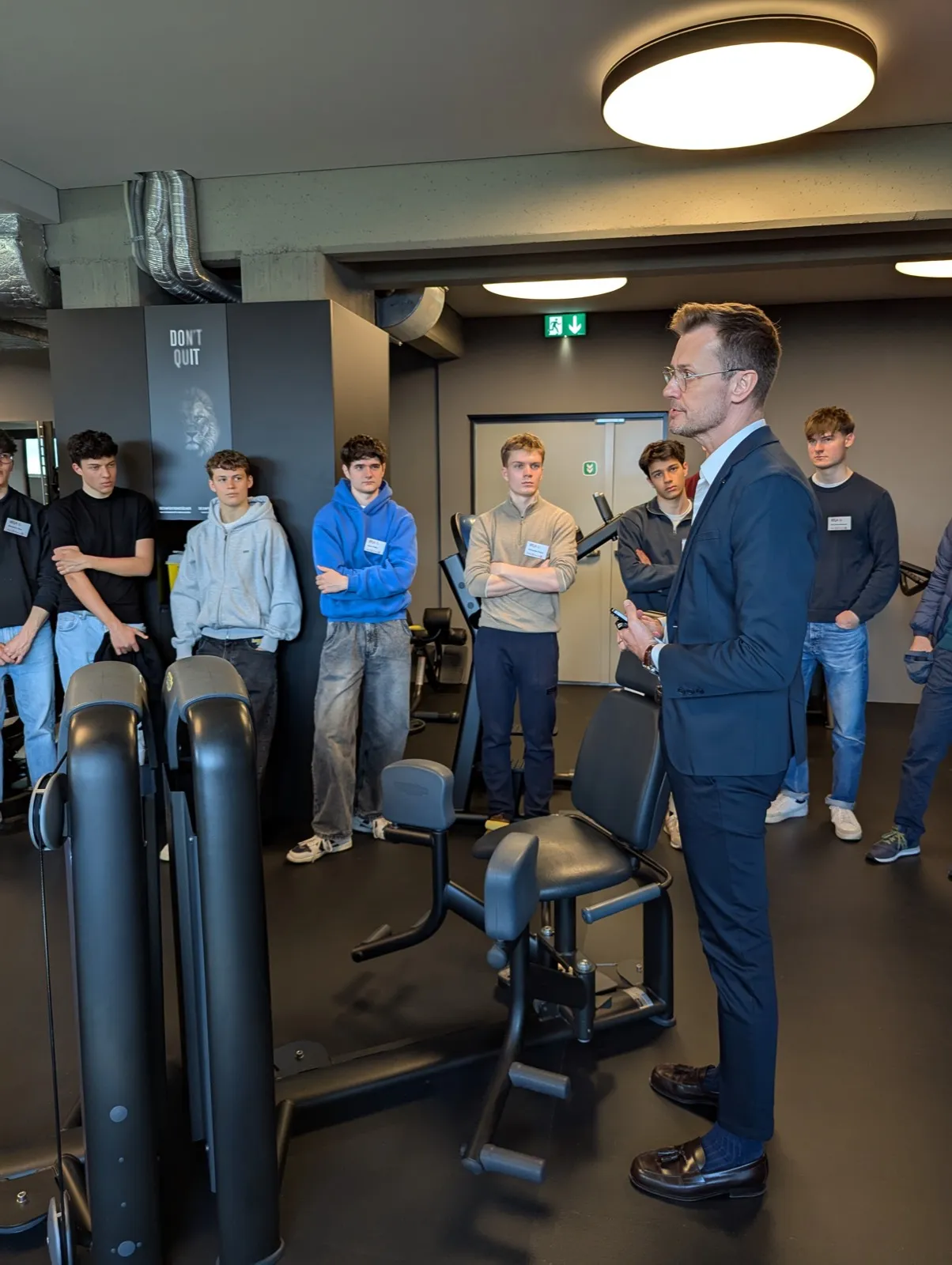 A man in a dark blue suit addressing a group of young men standing around fitness equipment inside a modern gym with concrete ceilings