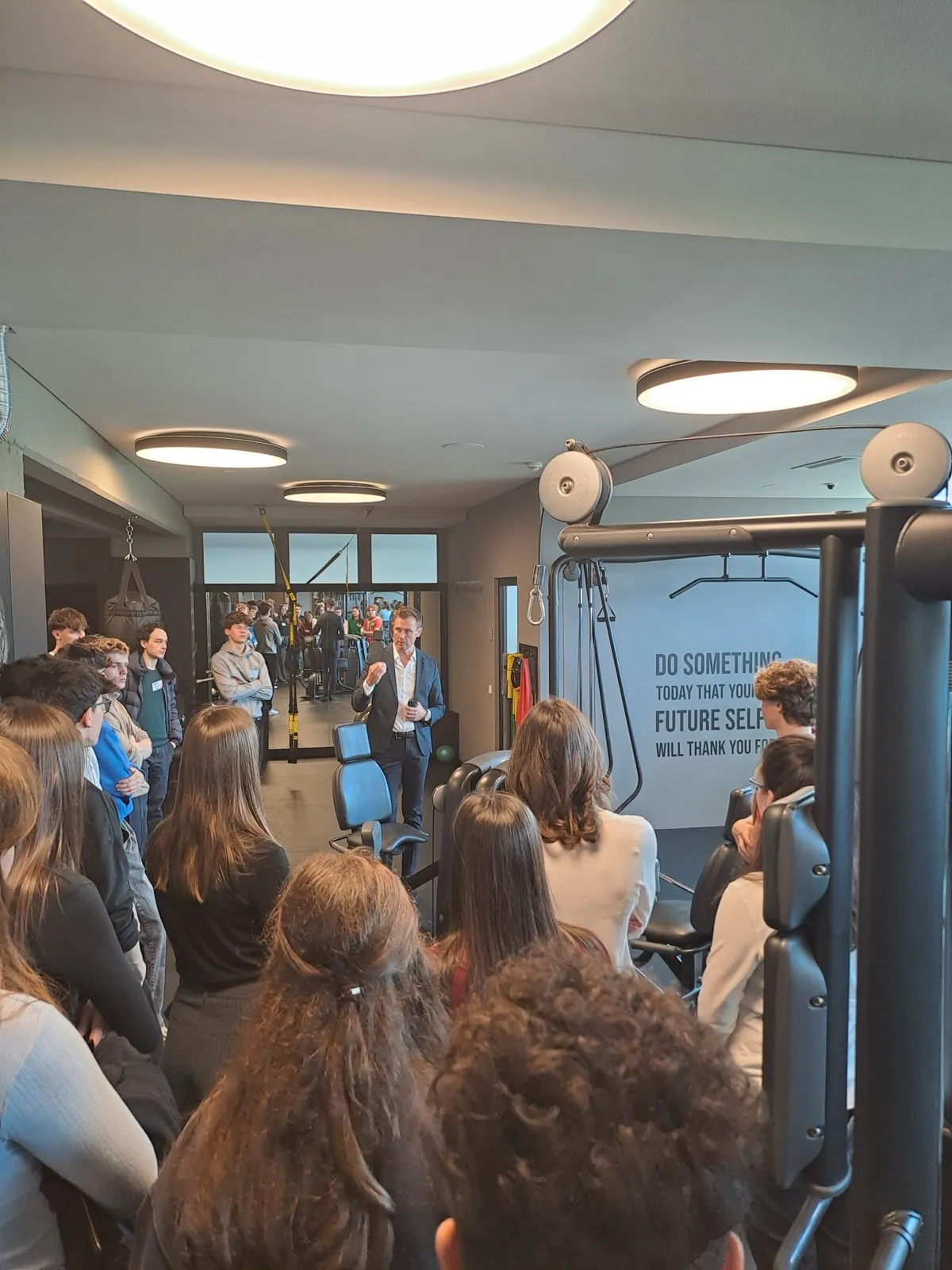 Rows of teenagers seated on wooden bleachers listening to a speaker beside gym cable machines, with a motivational wall quote in the background