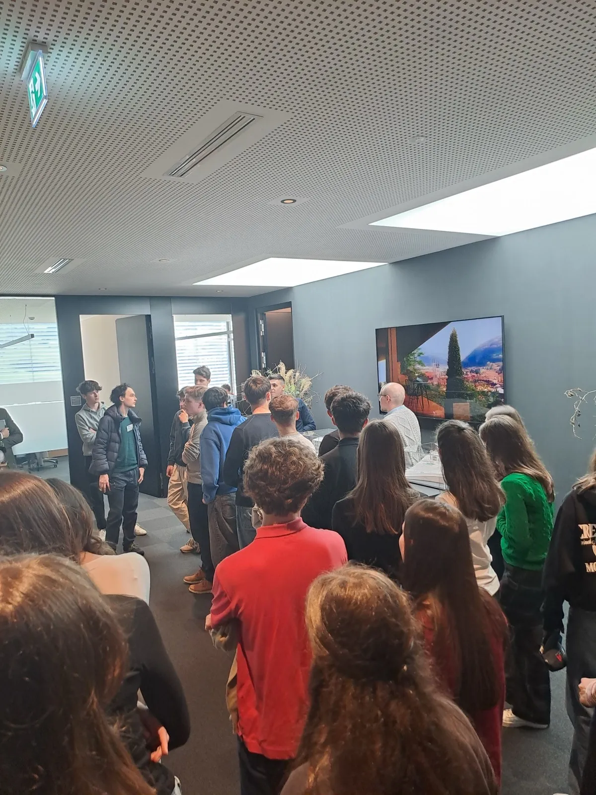 A crowd clustered in a bright hall watching a wall-mounted screen that shows a mountain landscape, some wearing red jackets and lanyards