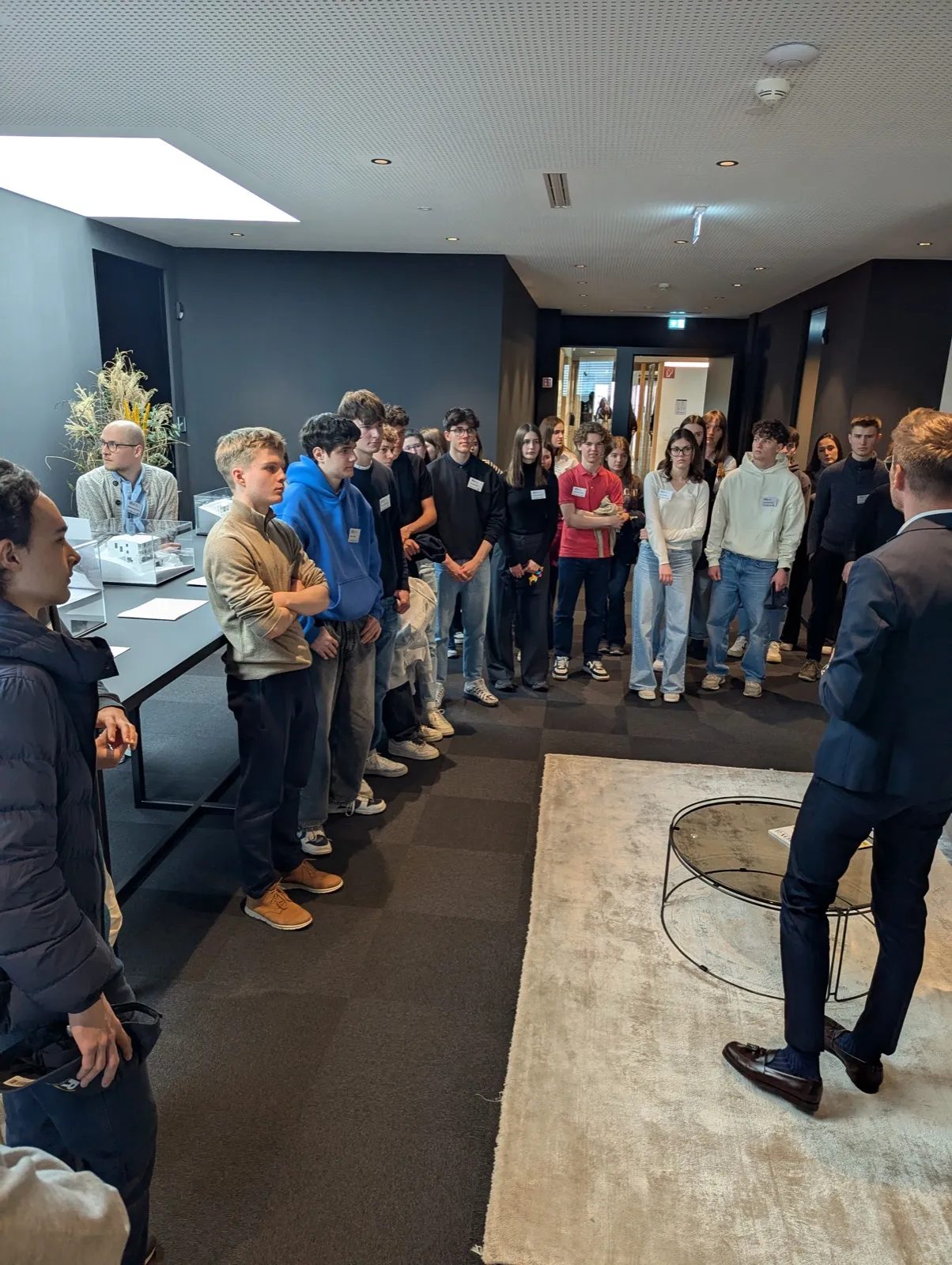 A speaker in a suit addressing about twenty teenagers standing in a dark-walled showroom next to a table with an architectural model