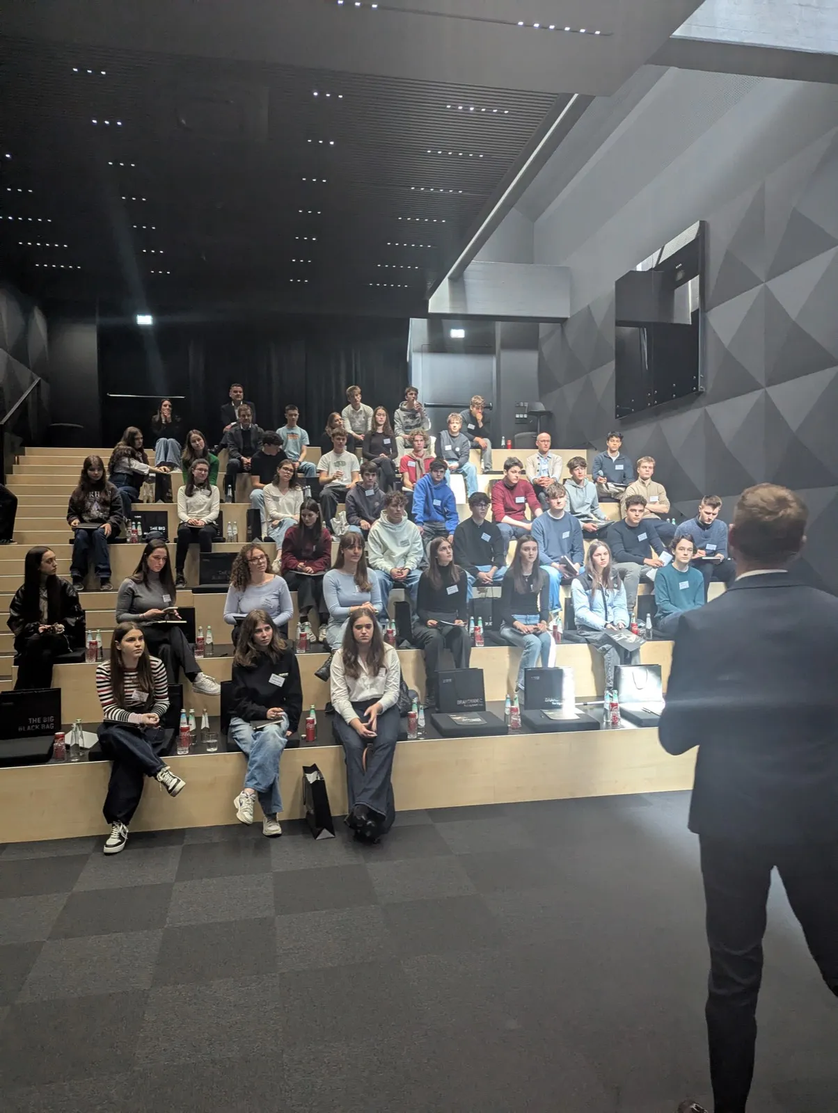 Speaker viewed from behind gesturing toward a full auditorium of young people seated on stepped wooden benches with water bottles and bags