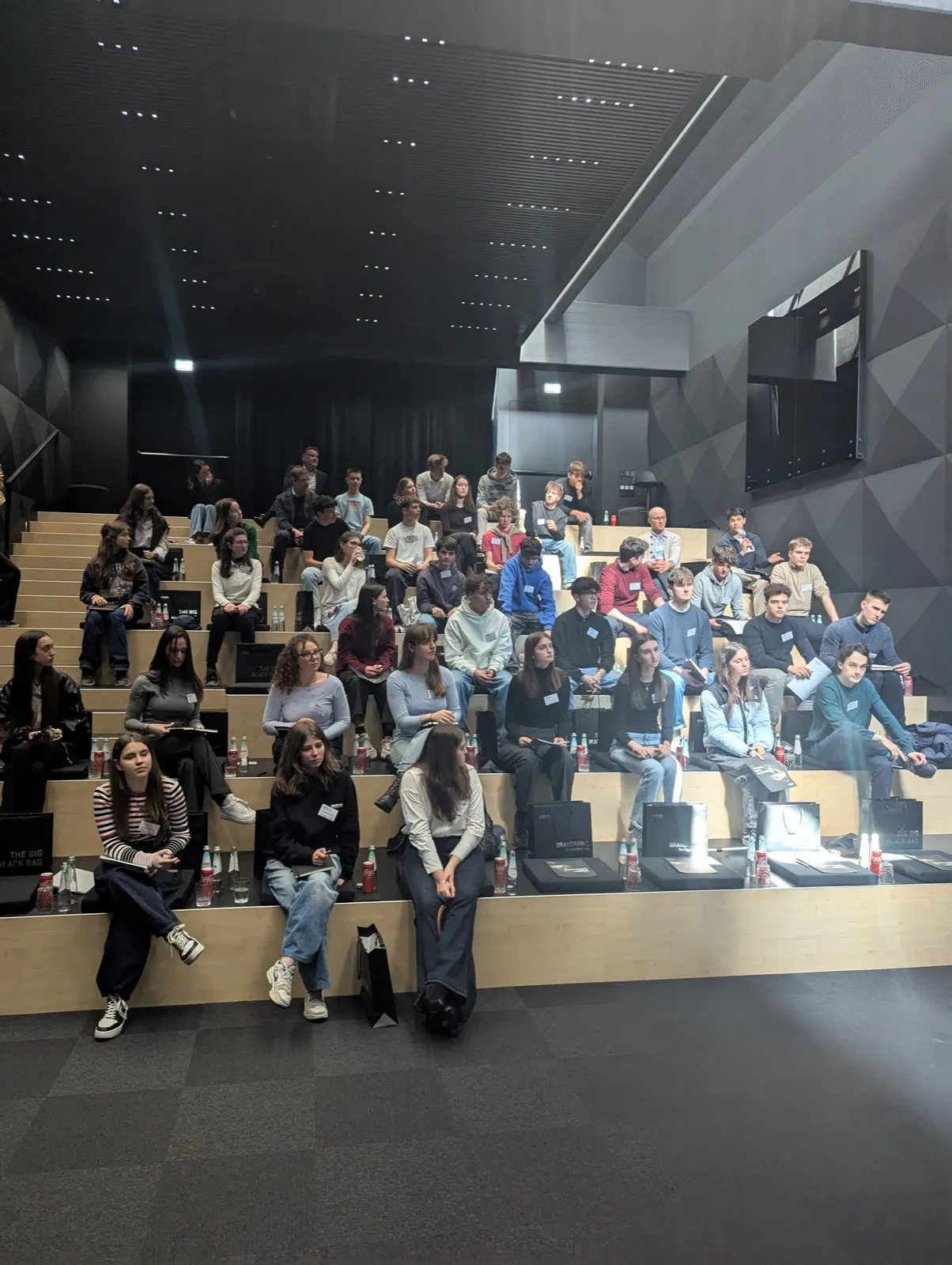 Wide auditorium view of about forty youth participants scattered across tiered wooden platforms, each spot marked with black gift bags and beverages