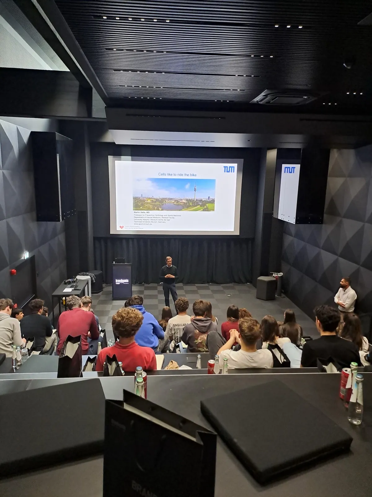 A presenter standing beside a projected slide about carbohydrates and cycling while participants take notes at long conference tables