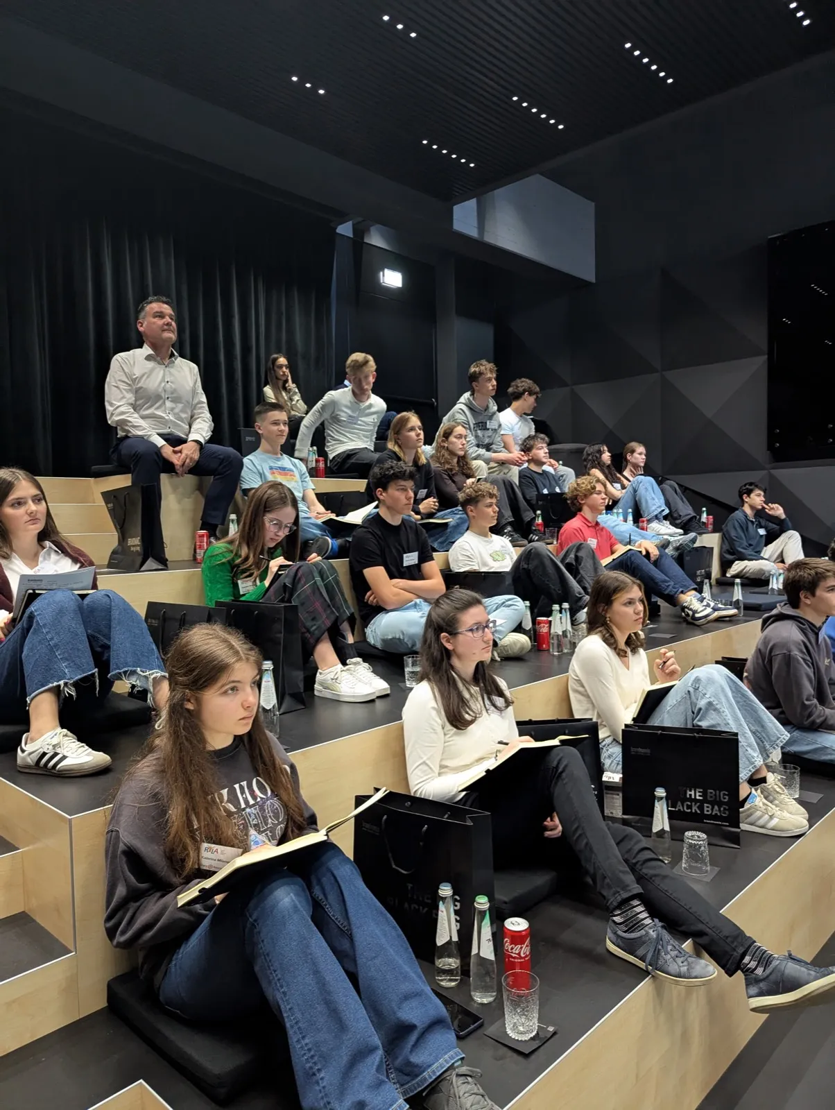 A girl in a printed sweatshirt writing in a notebook while surrounded by peers on tiered seating, water bottles and a red soft drink can nearby