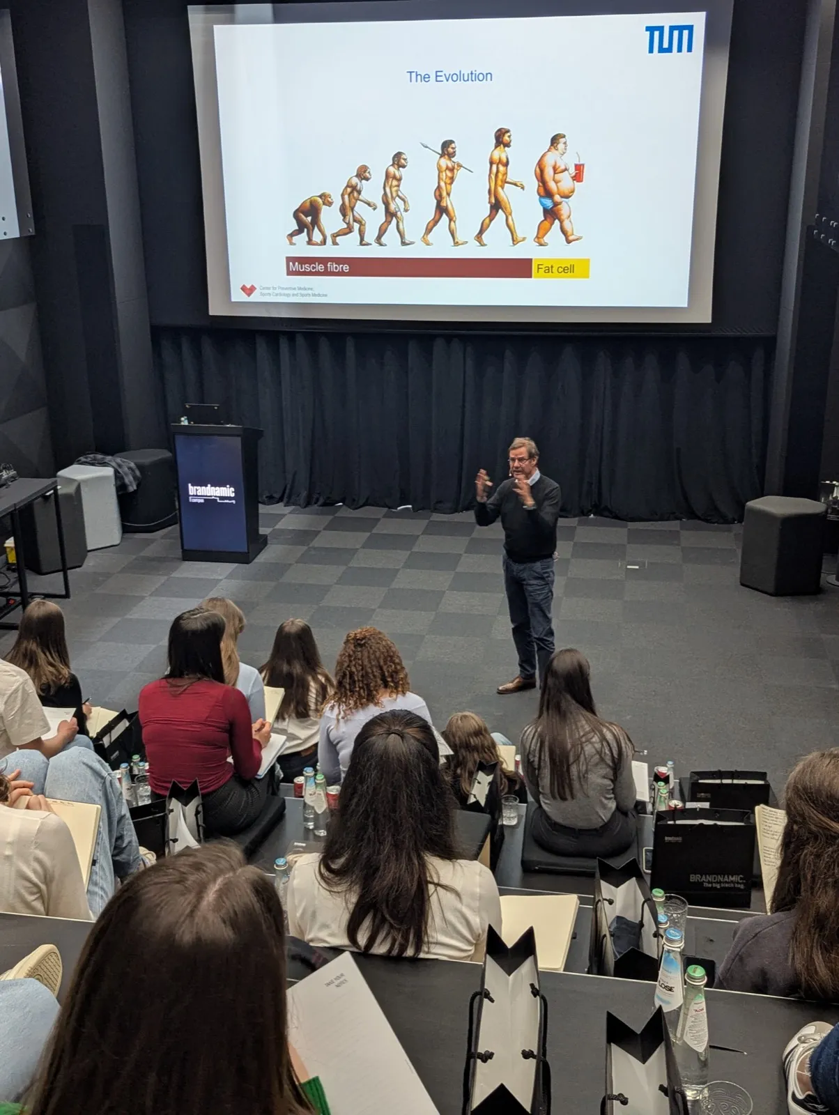 An instructor gesturing in front of a wide screen showing a human evolution illustration labelled muscle fibre and fat cell, seated audience taking notes
