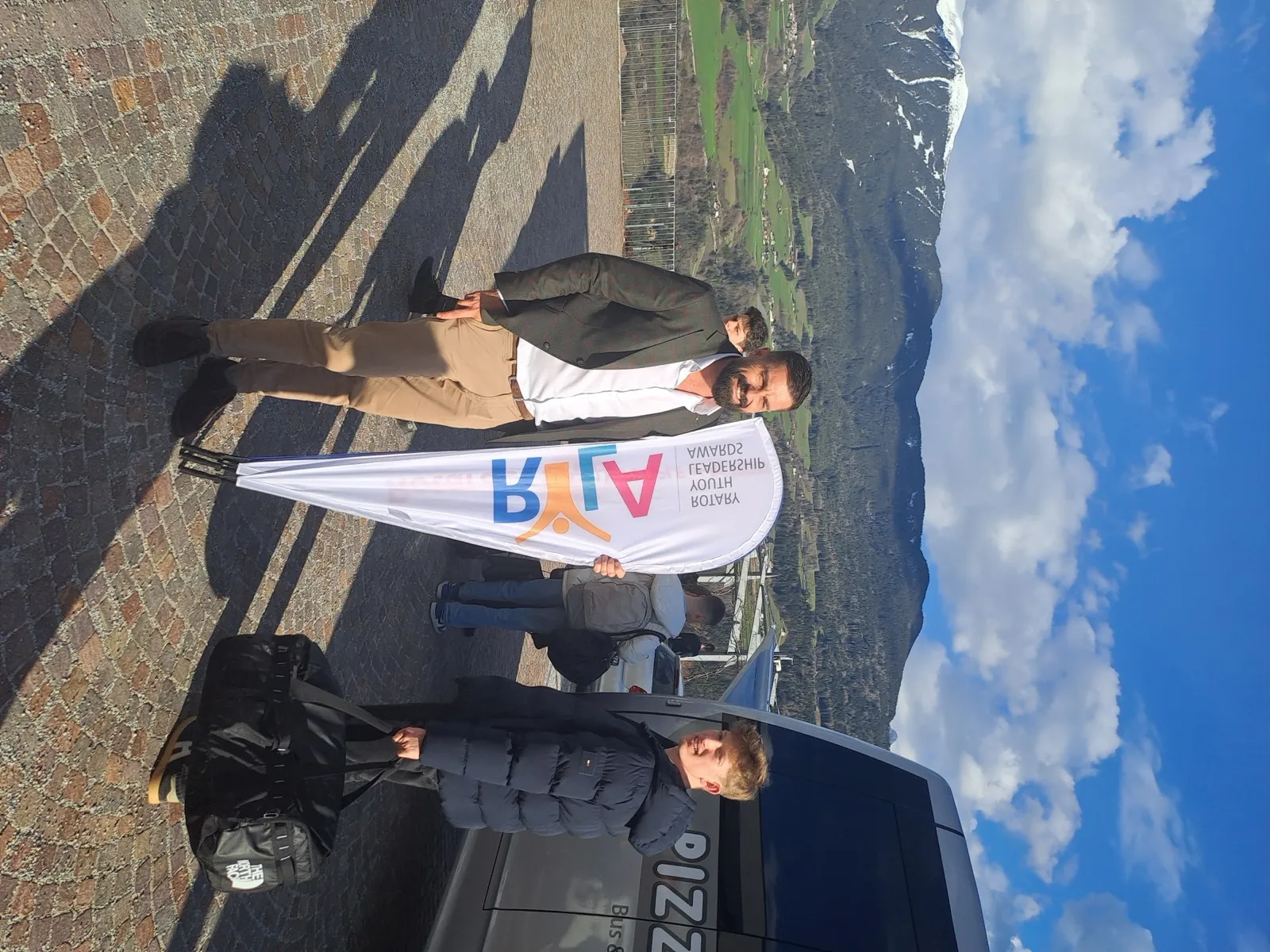 Two people posing on a stone plaza beside a Rotary Youth teardrop banner with snowy mountain peaks and a minibus behind them