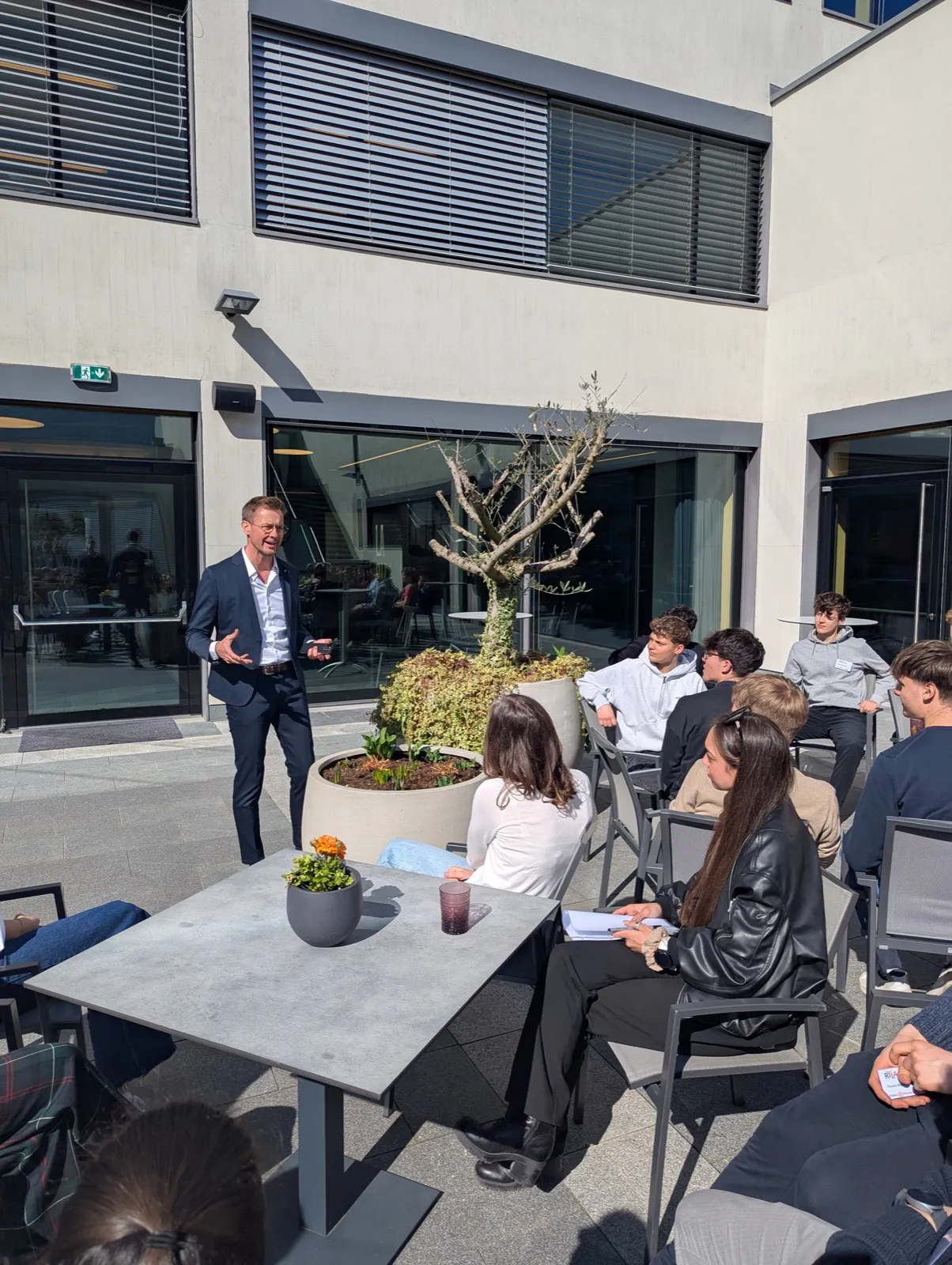 A speaker in a navy blazer talking to seated teenagers around a sunny outdoor terrace table with a small potted olive tree between them