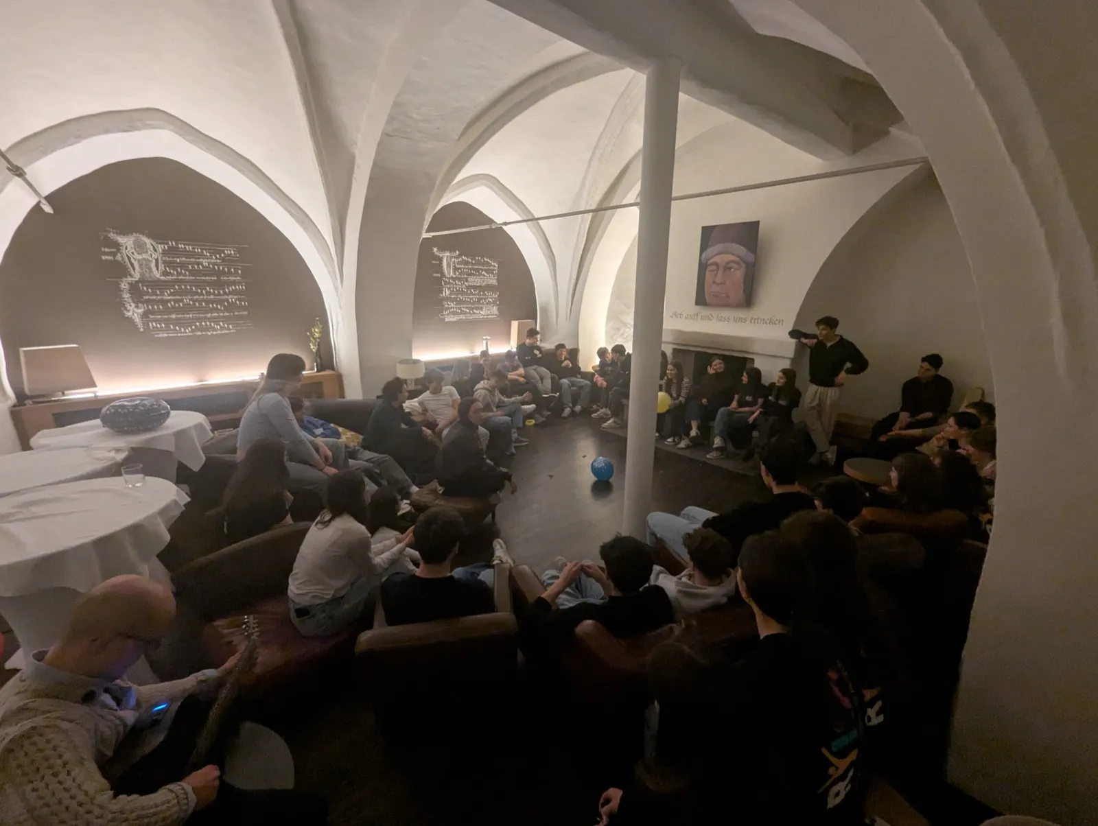 A large group lounging on sofas beneath vaulted white monastery arches decorated with musical notation projections and a portrait bearing an inscription