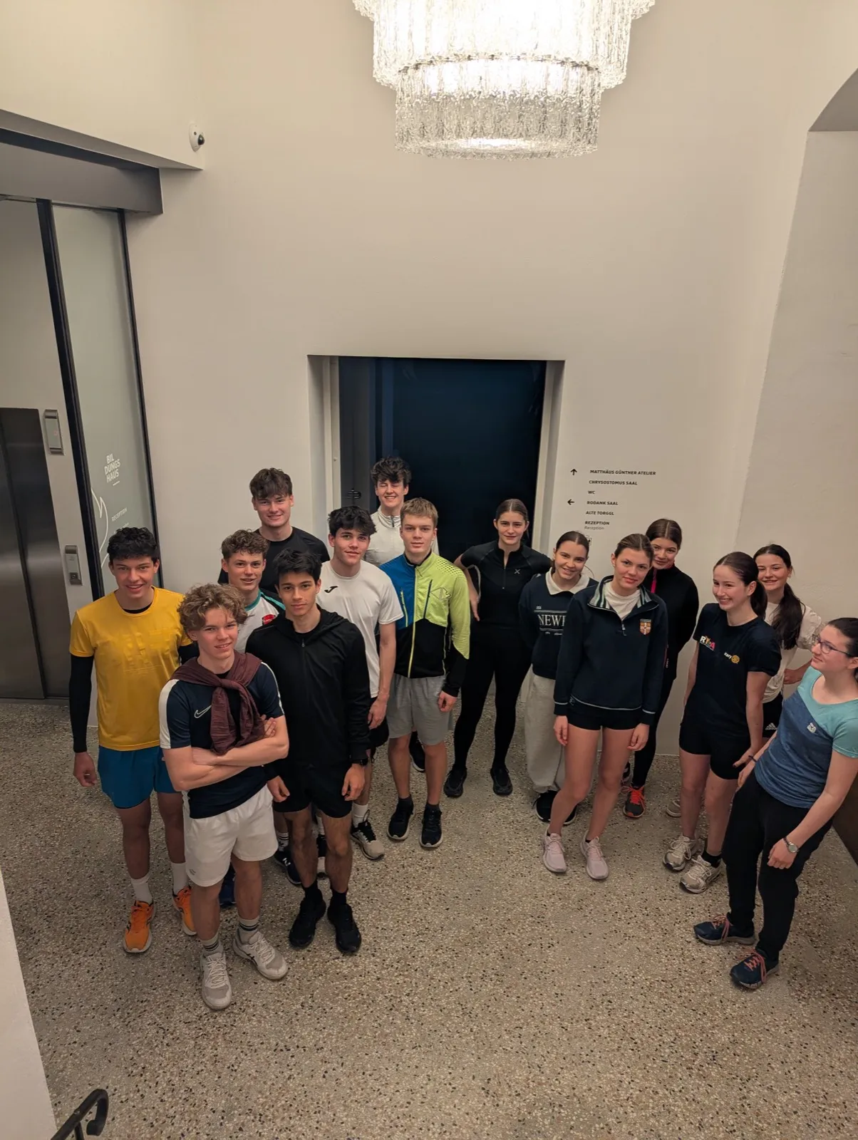 Group portrait of fifteen teenagers in sportswear standing under a large crystal chandelier in a hotel lobby beside an elevator and directional signage