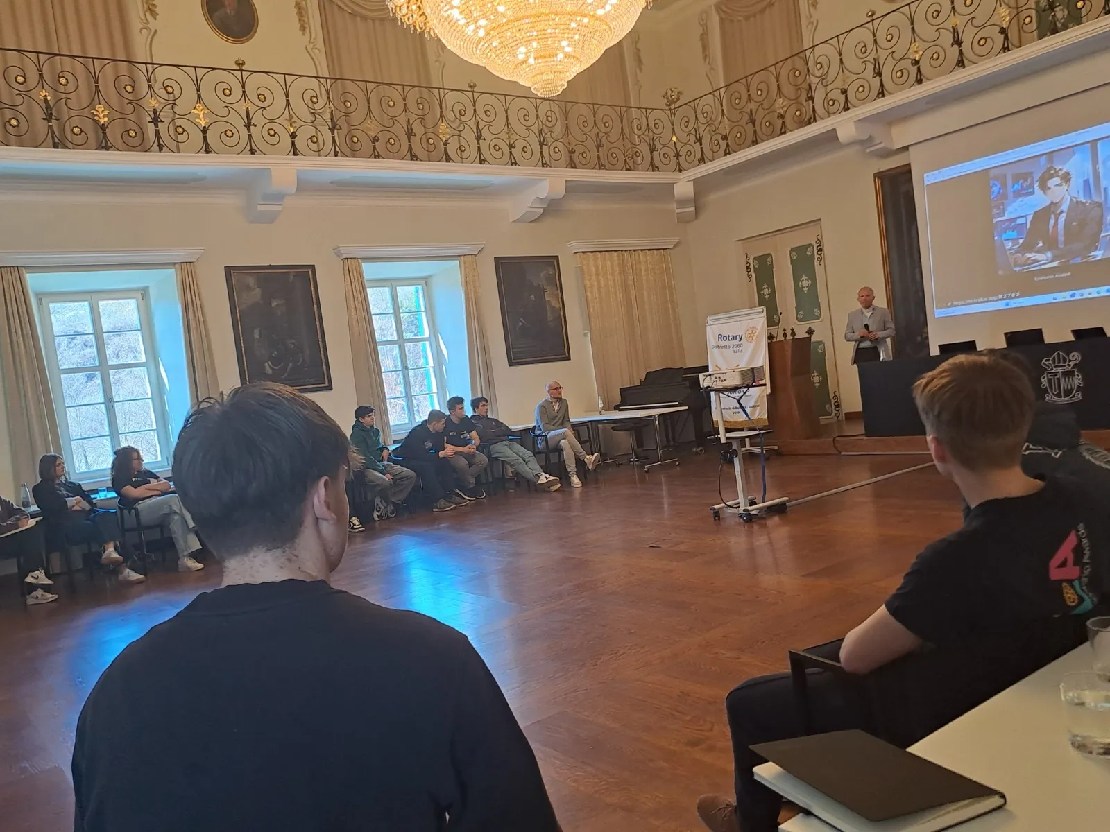 An ornate baroque hall with chandelier and gilded balcony where teenagers watch a projected interview beside a Rotary Youth rollup banner