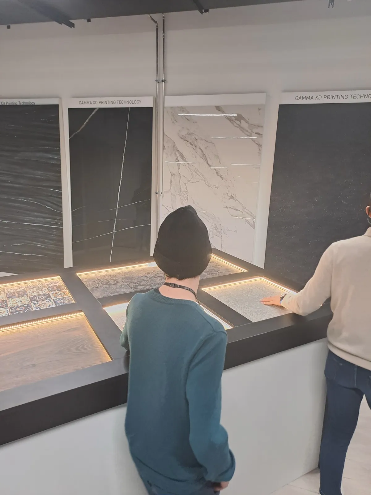 Two visitors in a tile showroom examining large marble-look wall panels illuminated by warm backlighting, one wearing a beanie and a teal sweater