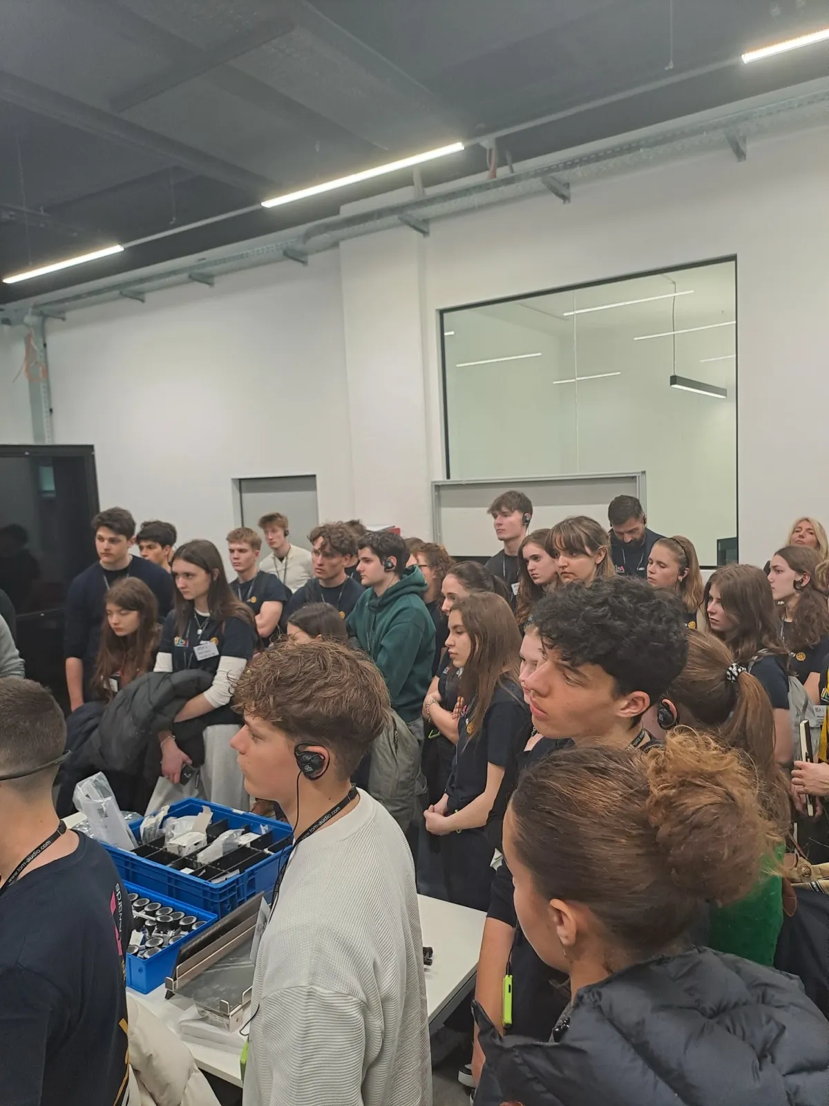 A densely packed crowd of youths watching a technician in a white shirt operate equipment out of a blue crate near a glass staircase