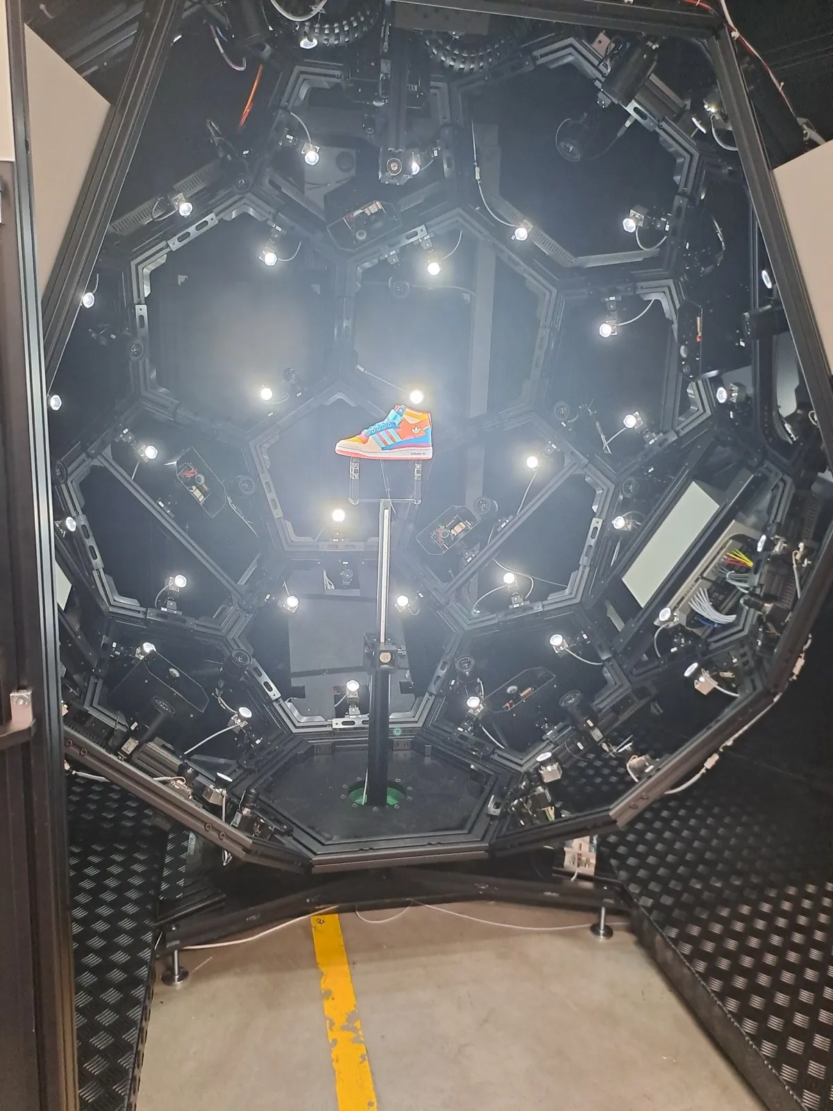 Top-down view into a black octagonal photogrammetry dome with ring lights aimed at a colourful sneaker suspended above the central platform